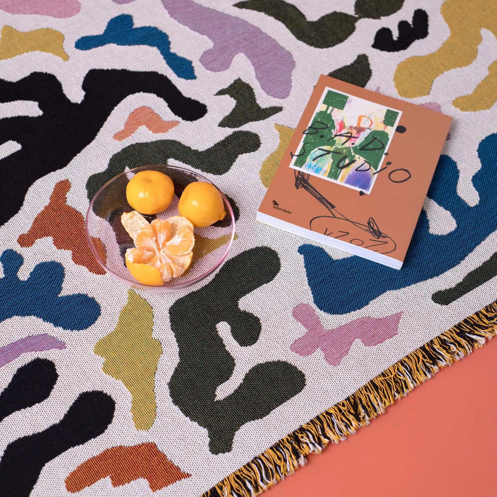The Slowdown Studio Mercer Throw, featuring a vibrant abstract pattern and made from recycled cotton, serves as a backdrop for a glass bowl of tangerines and an illustrated book resting on the fringed rug.