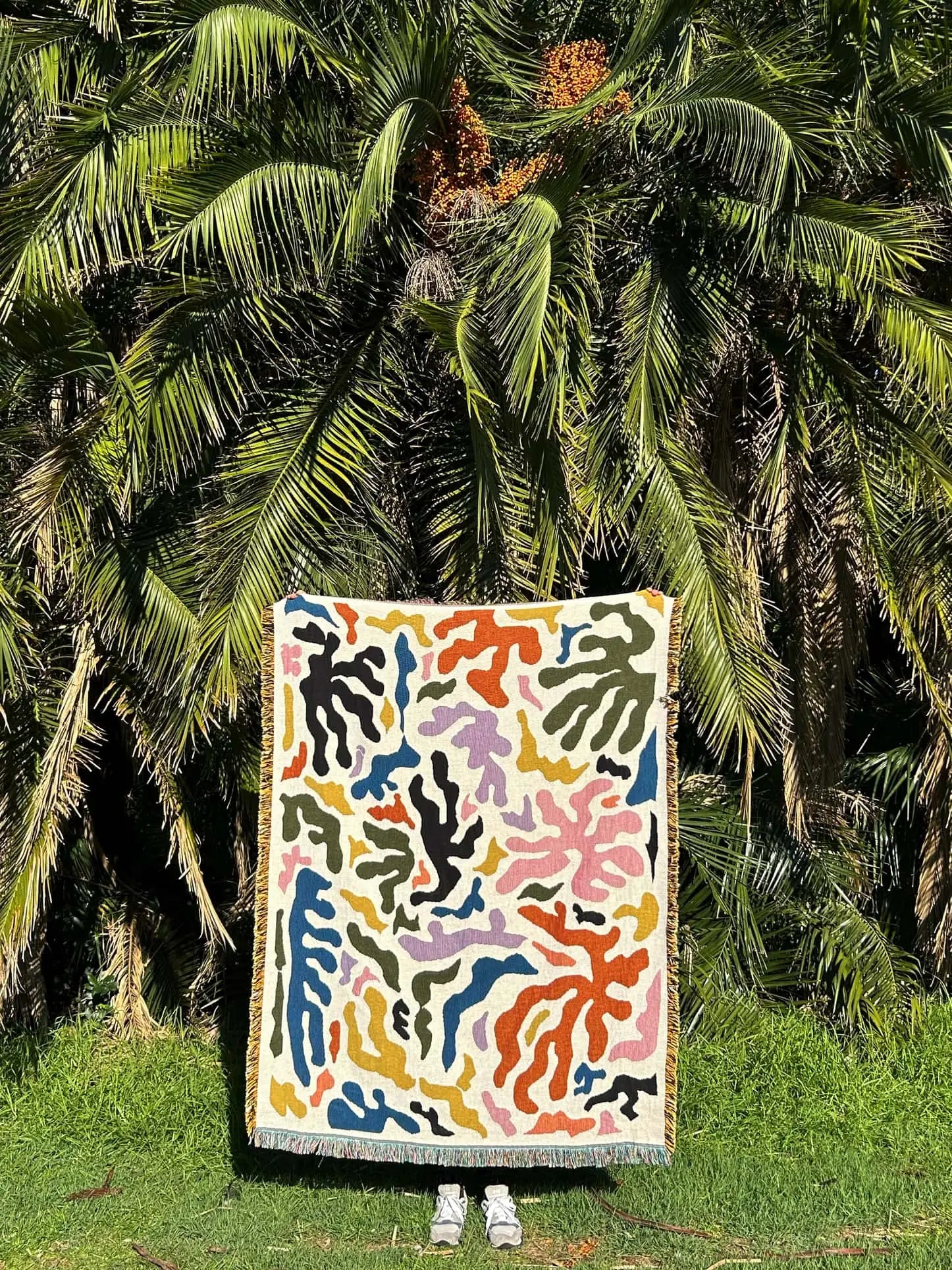 A person stands on grass before palm trees, holding up a colorful abstract-patterned Slowdown Studio Mercer Throw made from recycled cotton, with just their shoes peeking out below the blanket.