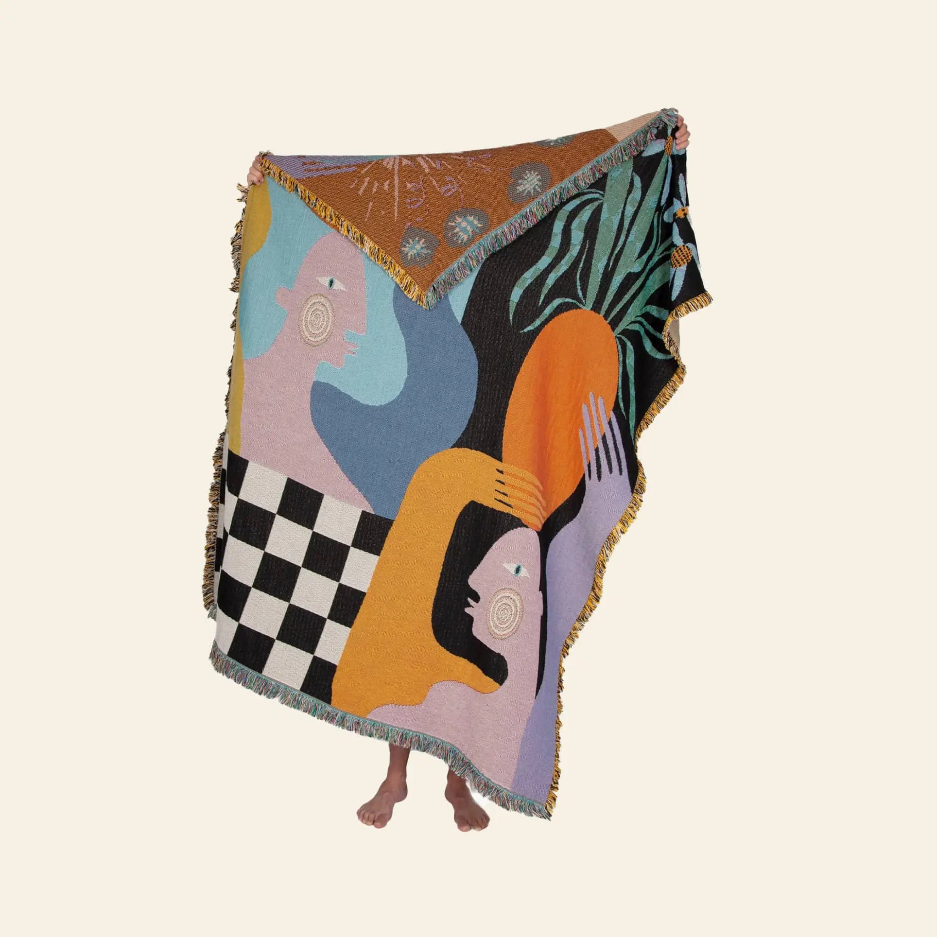 The Slowdown Studio Moretti Throw by Slowdown Studio is an eco-friendly woven blanket made from recycled cotton, featuring abstract figures, geometric patterns, and botanicals—shown held up with only the holders feet visible.