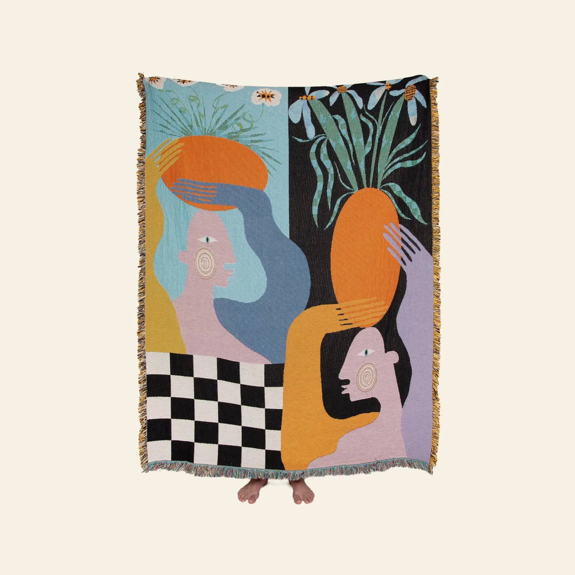 A person holds the Slowdown Studio Moretti Throw by Slowdown Studio, an eco-friendly tapestry made from recycled cotton with abstract colorful figures, large orange shapes, a checkerboard base, and leafy plants. Only their toes peek out.