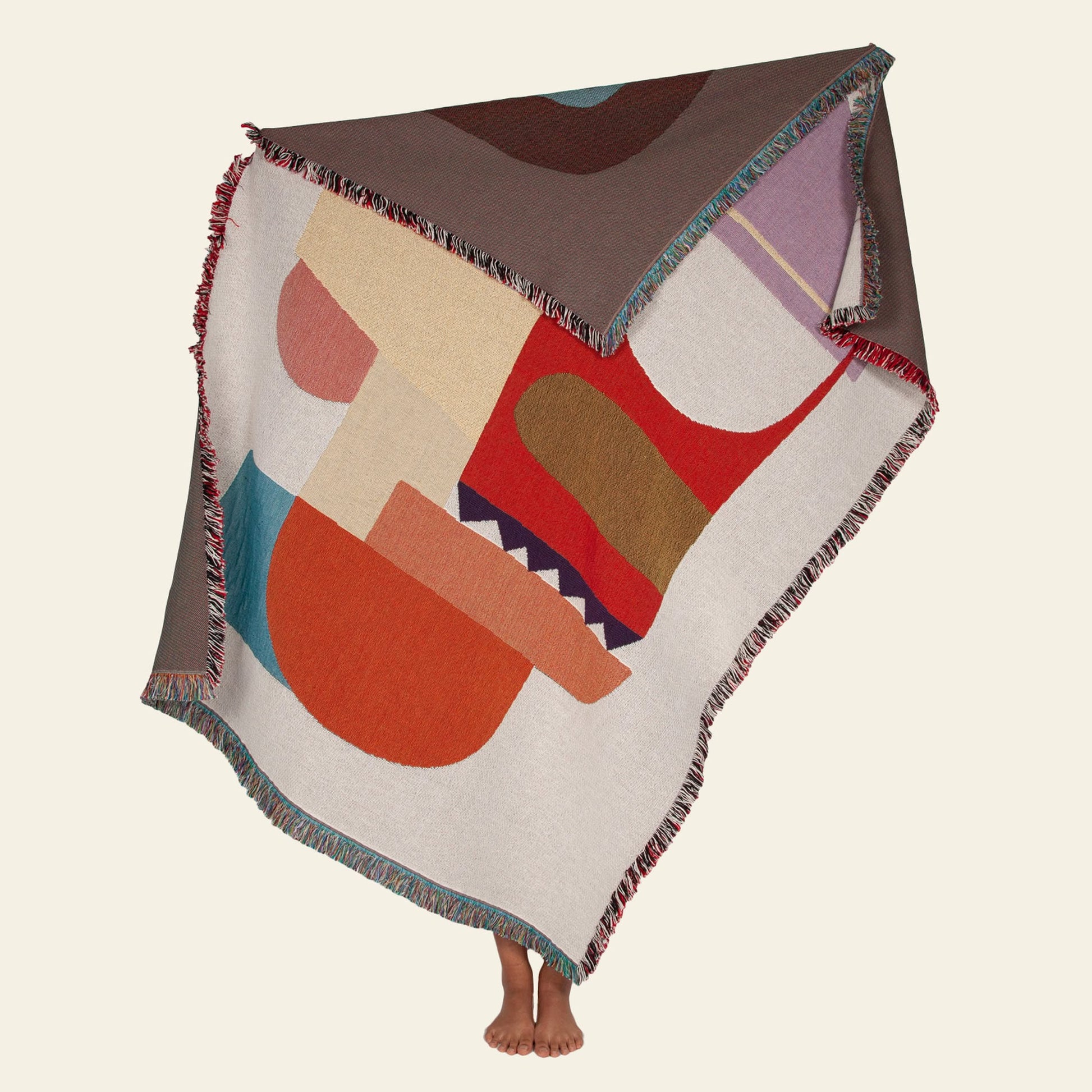A person with bare feet holds up the Slowdown Studio Oroza Throw—a large, fringed blanket by Slowdown Studio featuring an abstract, colorful geometric design—made from eco-friendly recycled cotton, set against a plain background.