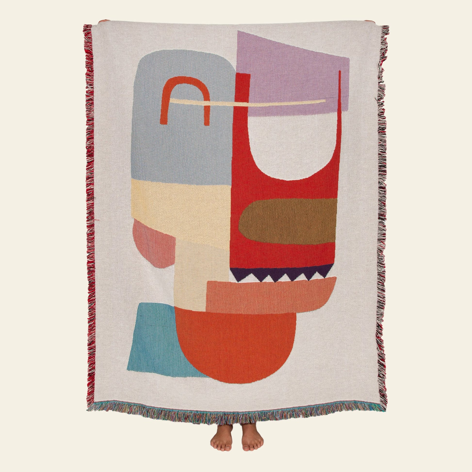 A person holds up the Slowdown Studio Oroza Throw, a large, colorful woven blanket by Slowdown Studio made from recycled cotton, showing its abstract geometric design with their feet visible below the edge.