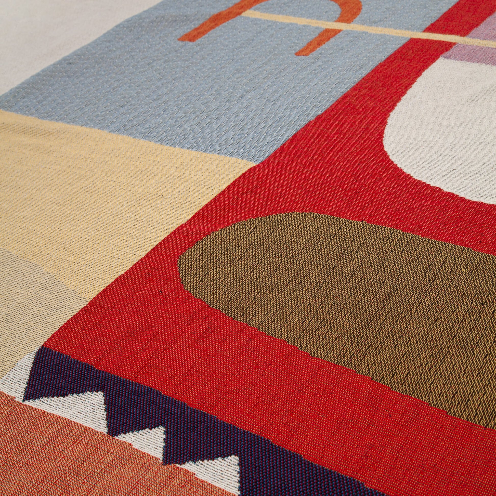 A close-up of the Slowdown Studio Oroza Throw, a colorful woven textile by Slowdown Studio made from recycled cotton, featuring geometric shapes in red, blue, beige, and olive green with zigzag and curved patterns.
