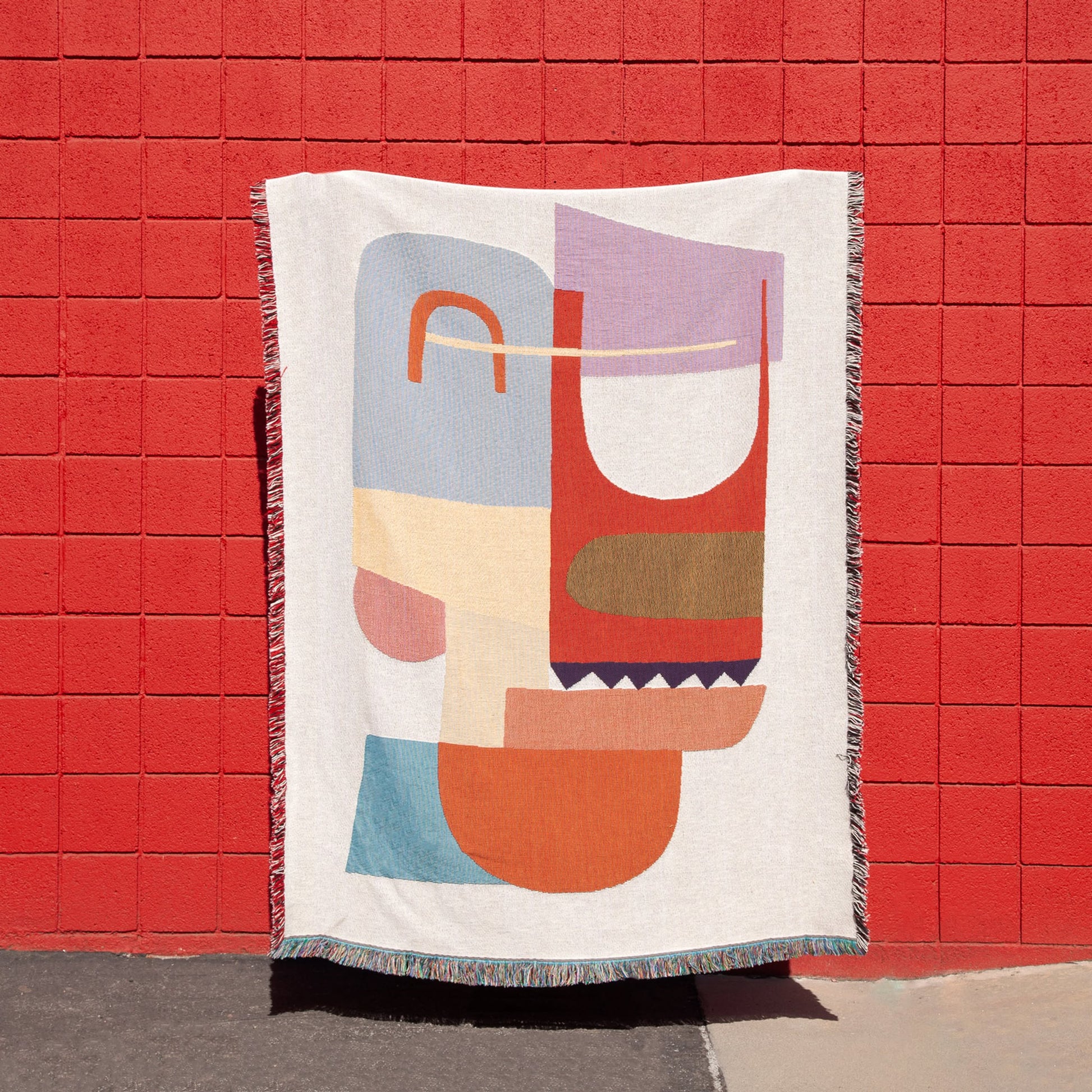 The Slowdown Studio Oroza Throw by Slowdown Studio, made from recycled cotton and featuring a colorful geometric pattern, hangs against a vivid red brick wall, with part of it casting a shadow on the sidewalk.