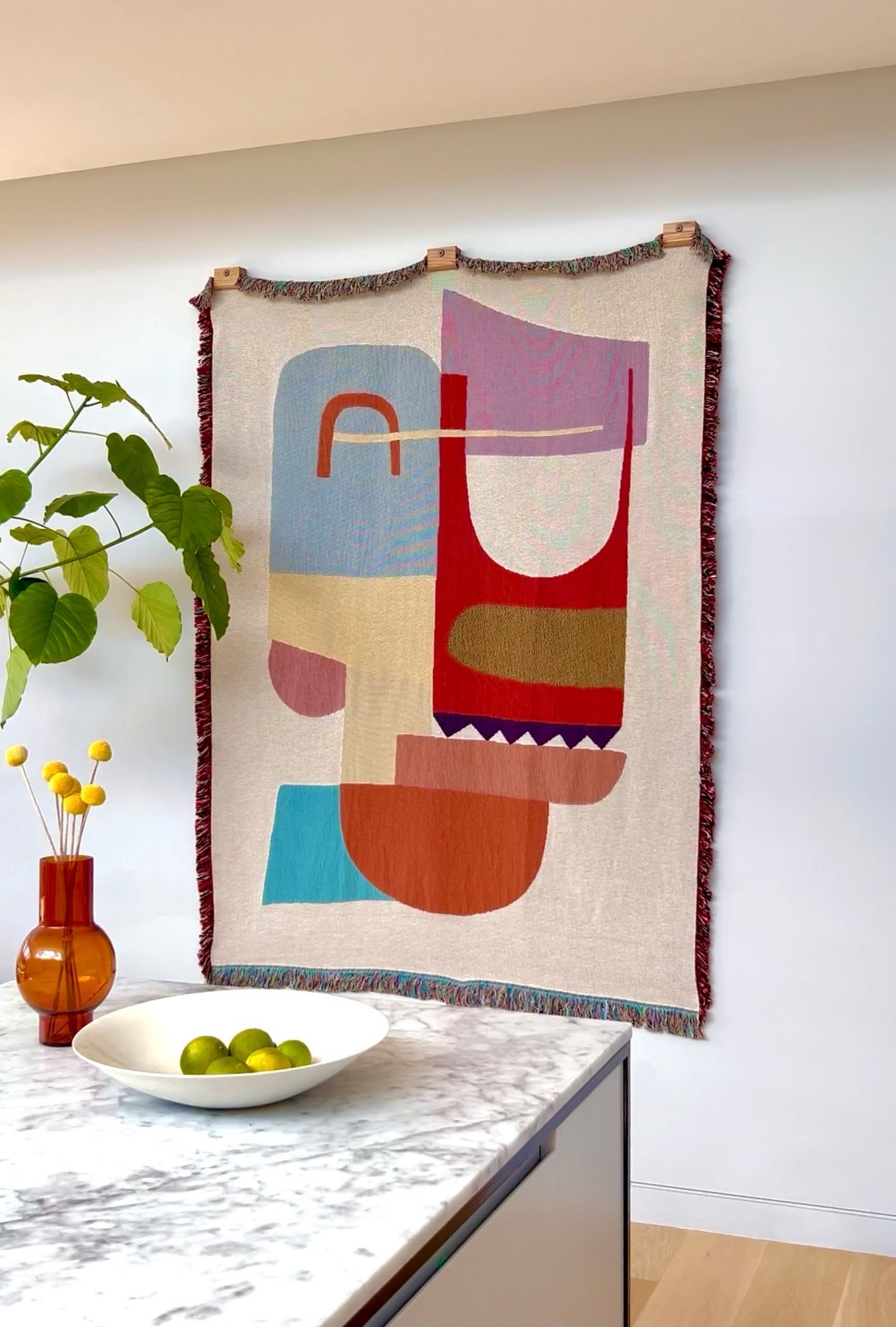 A vibrant abstract tapestry, the eco-friendly Slowdown Studio Oroza Throw crafted from recycled cotton, hangs above light wood floors. In front, a marble countertop displays a white bowl of green limes and a brown glass vase with yellow billy balls.