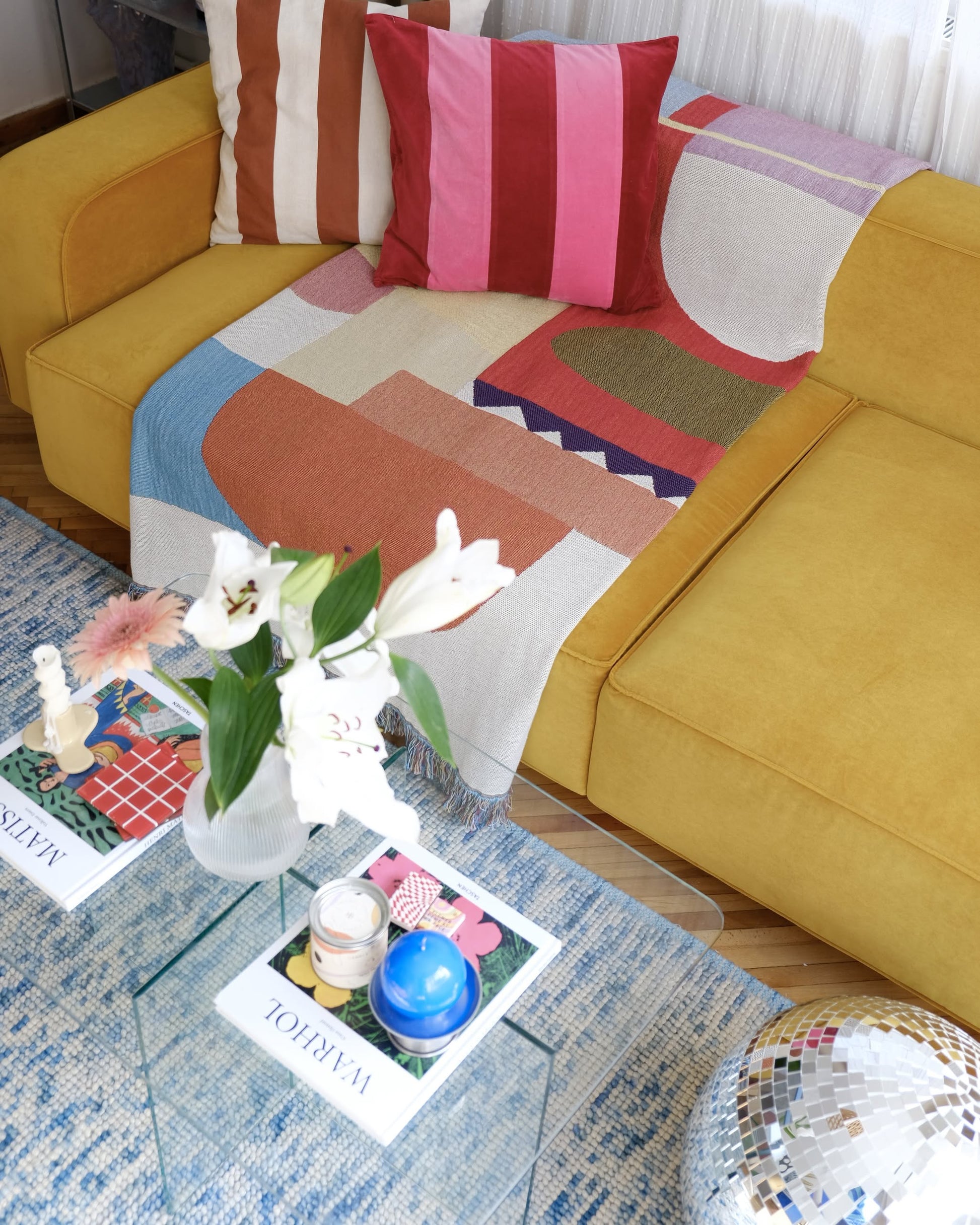 A yellow sofa features colorful, geometric pillows and the eco-friendly Slowdown Studio Oroza Throw made from recycled cotton. In front, a glass coffee table displays magazines, candles, a vase with white lilies, and a disco ball sits nearby.