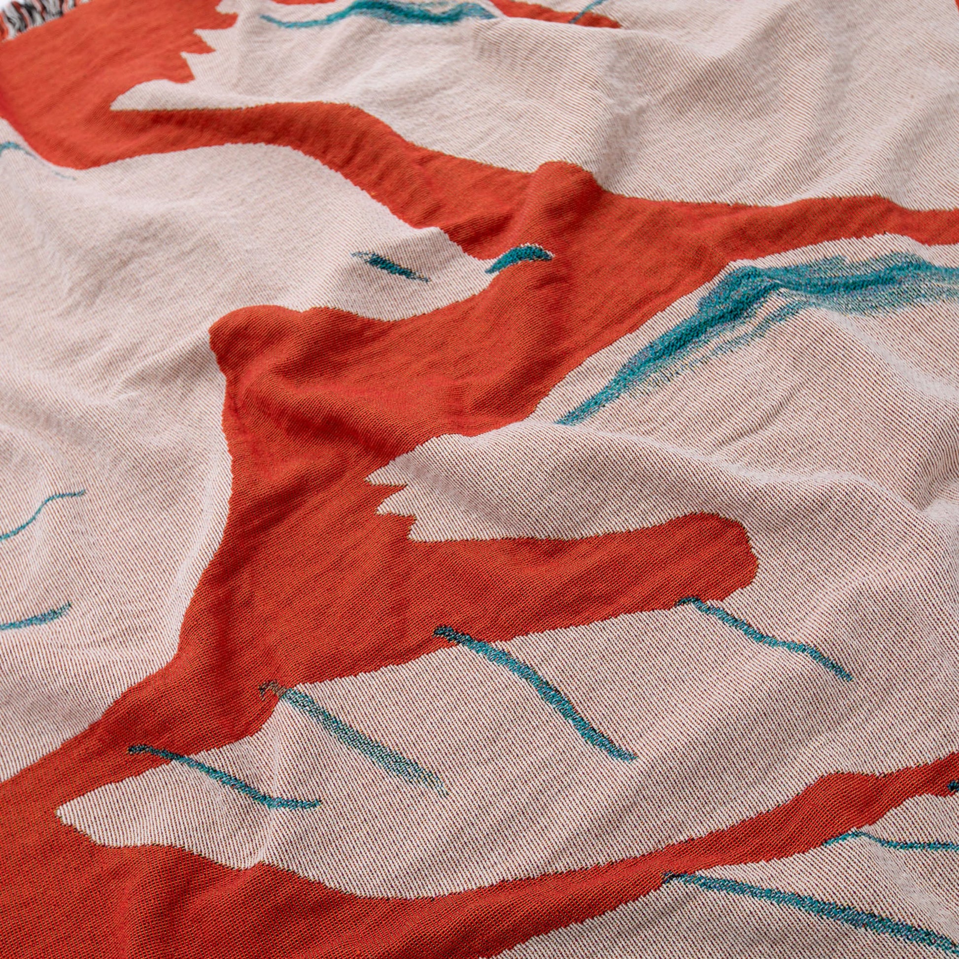 A close-up of the Slowdown Studio Passaro Throw by Slowdown Studio, crafted from recycled cotton with an abstract red, teal, and off-white pattern. The eco-friendly fabric features bold, irregular lines and shapes with a soft, textured finish.