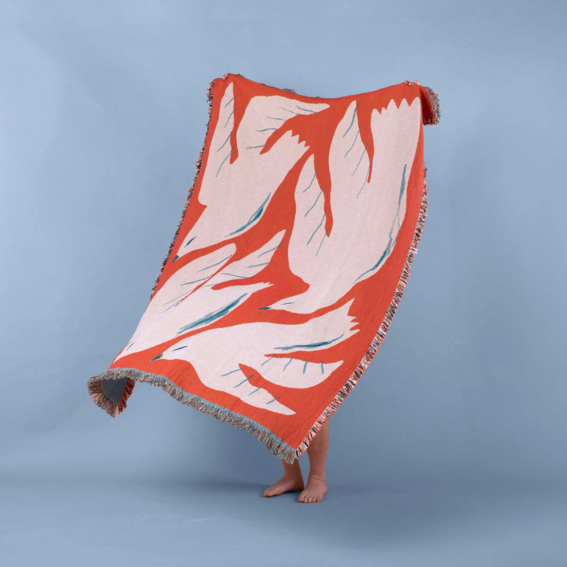 Standing barefoot on a light blue background, a person holds the Slowdown Studio Passaro Throw—an eco-friendly recycled cotton blanket from Slowdown Studio featuring an abstract orange, pink, and teal design. Only their feet show beneath the edge.
