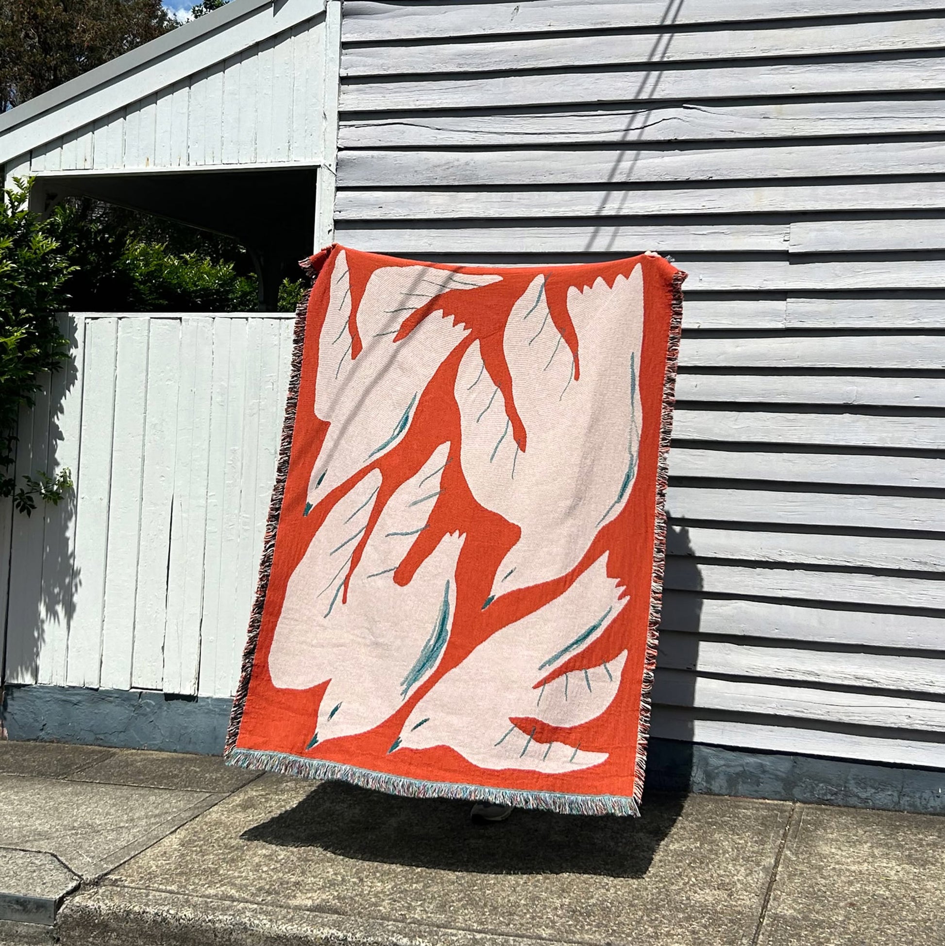 The Slowdown Studio Passaro Throw by Slowdown Studio is made from recycled cotton and features abstract red, white, and teal shapes. It hangs on a light gray wooden wall outside, casting a shadow on the sidewalk in bright sunlight.