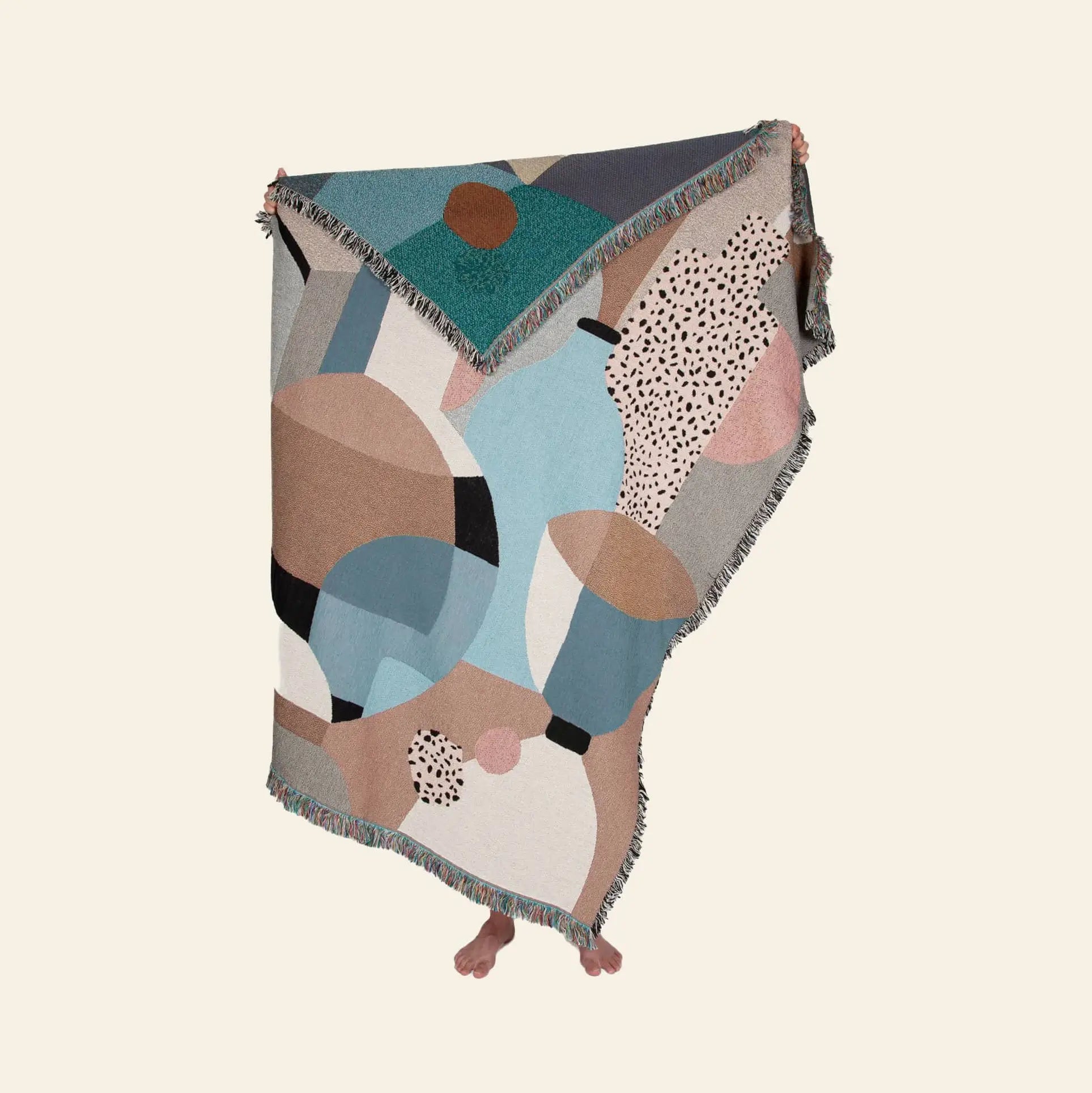A person stands covered in the Slowdown Studio Pollard Throw, an eco-friendly blanket made from recycled cotton with abstract geometric patterns in blue, tan, black, white, and pink. Only their feet show against a plain beige background.