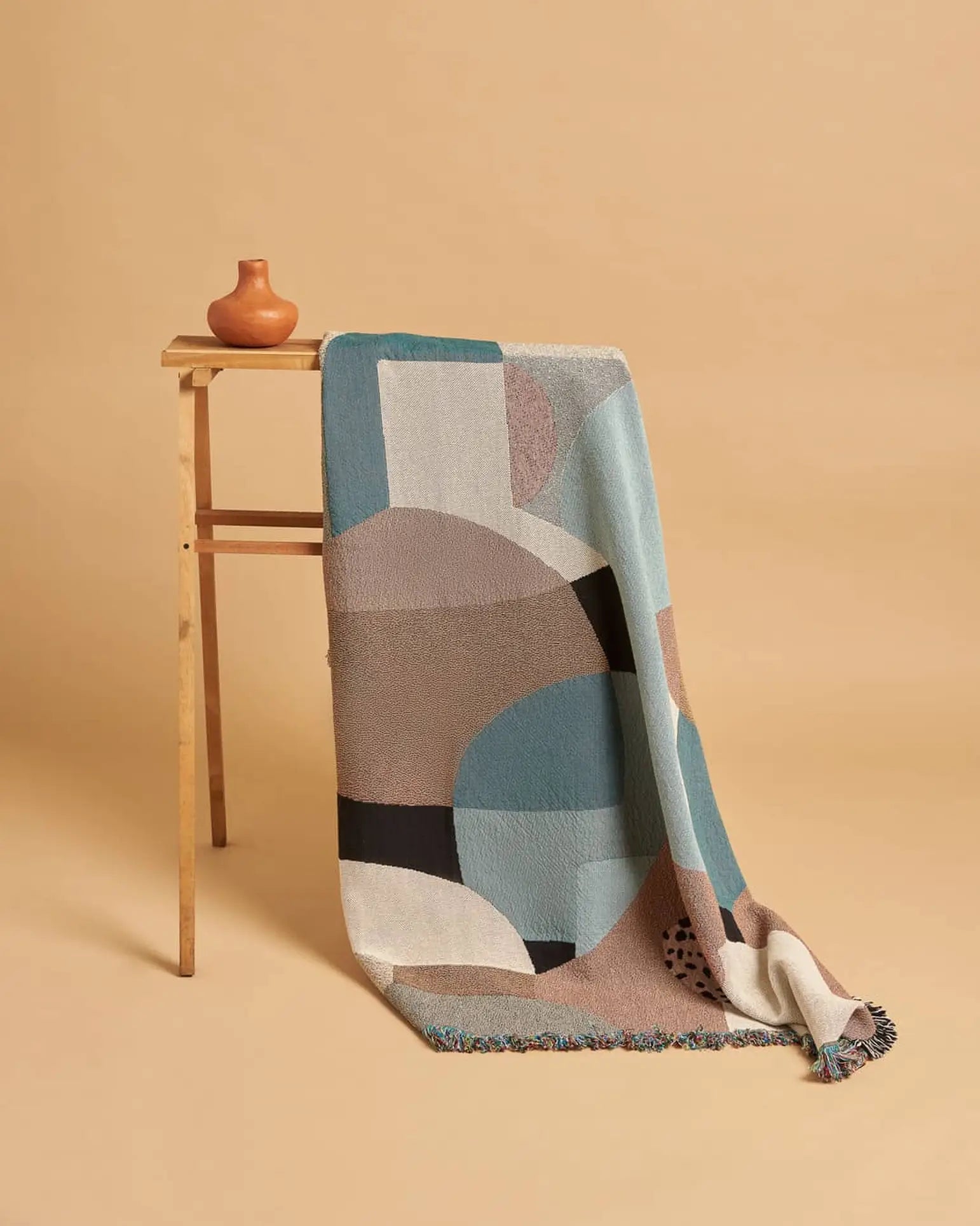 A wooden stand displays the Slowdown Studio Pollard Throw—a recycled cotton blanket in blue, beige, brown, and black—paired with a small round brown vase against a beige background. Eco-friendly by Slowdown Studio.