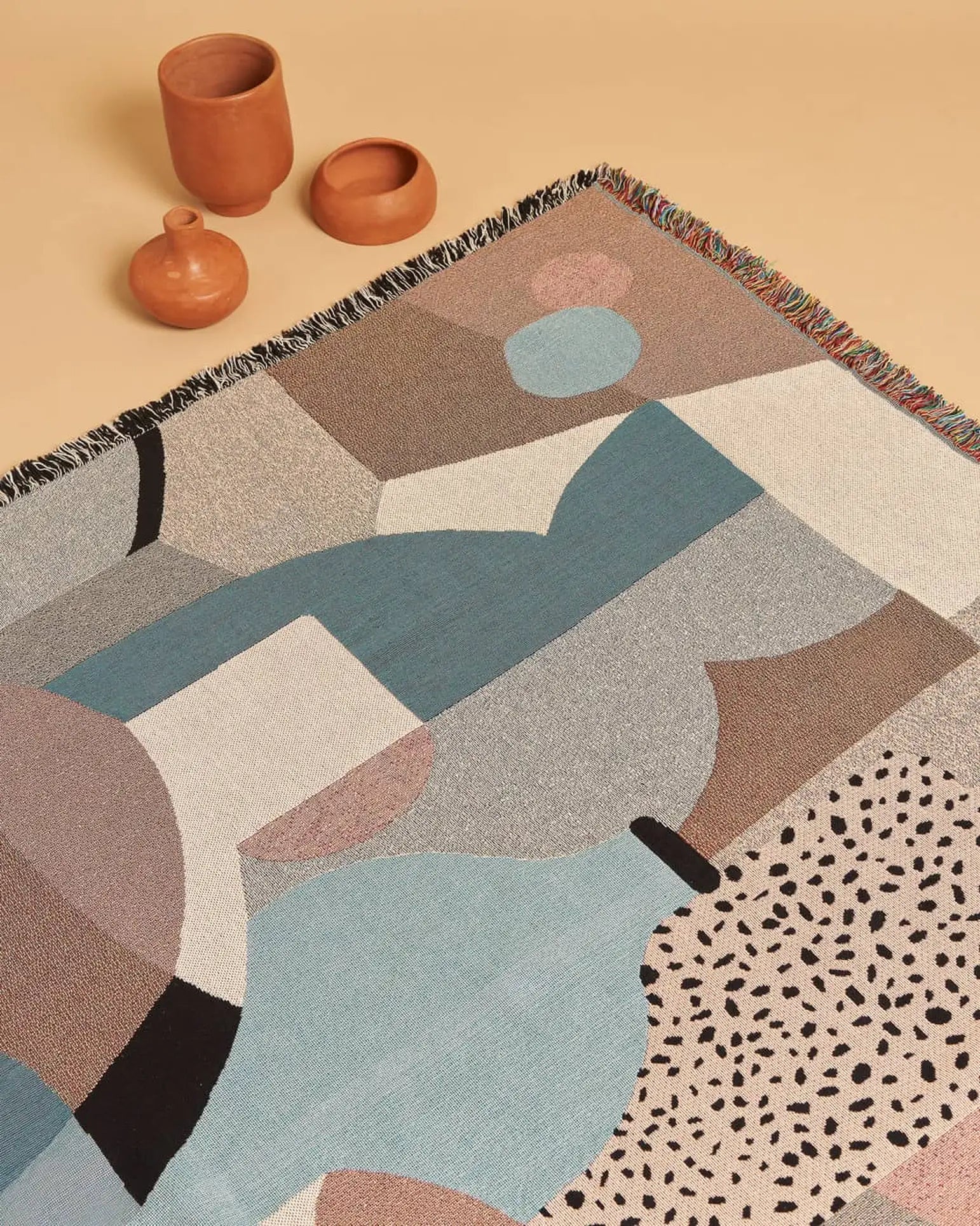 The Slowdown Studio Pollard Throw by Slowdown Studio, featuring a geometric pattern in blue, beige, brown, and black recycled cotton, is displayed on a light tan surface beside three small terracotta vessels of different shapes.