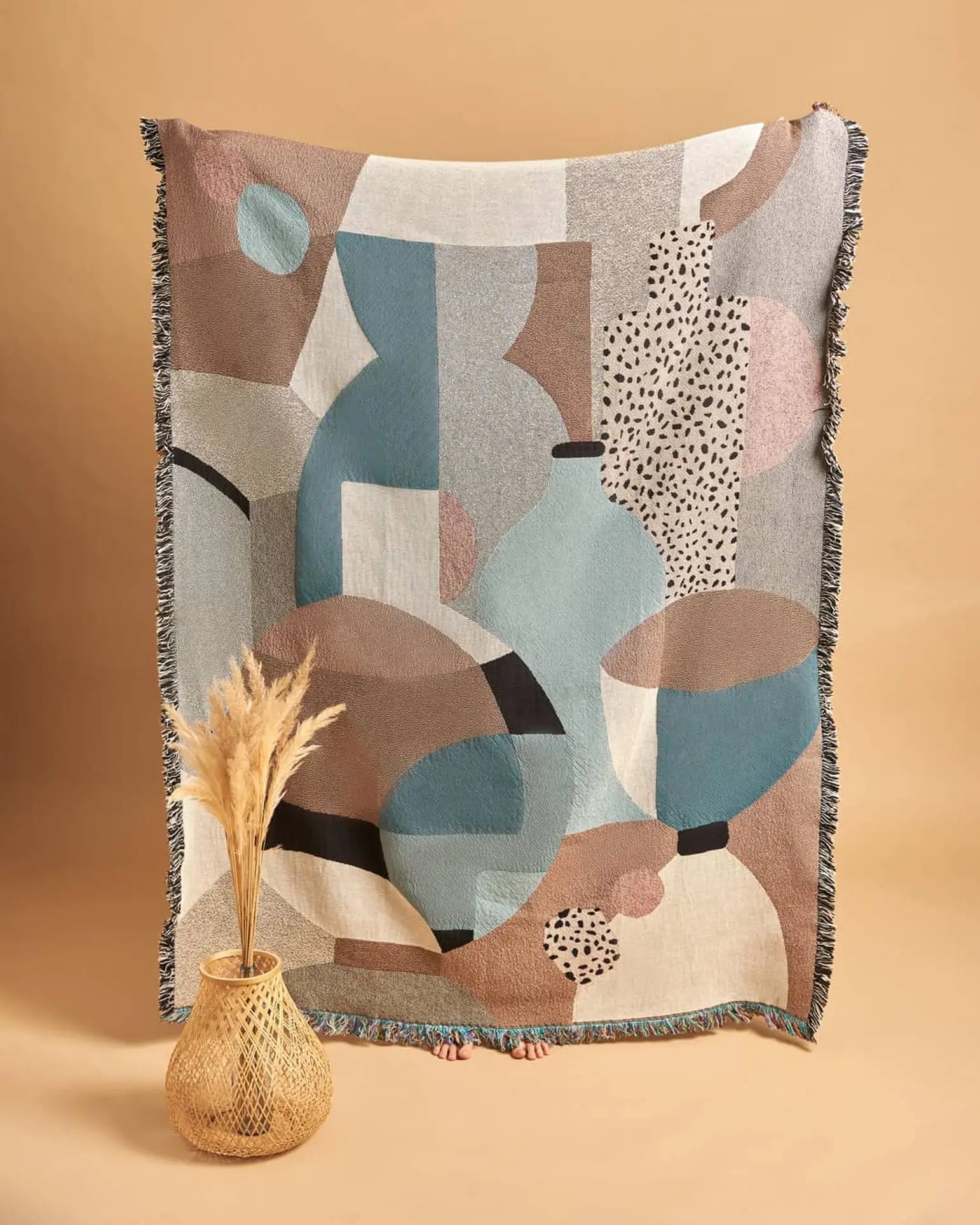 A person holds up the Slowdown Studio Pollard Throw, a large recycled cotton blanket in muted blue, brown, and beige hues. In front, an eco-friendly woven vase with pampas grass sits against a beige background. Only toes peek beneath the throw.