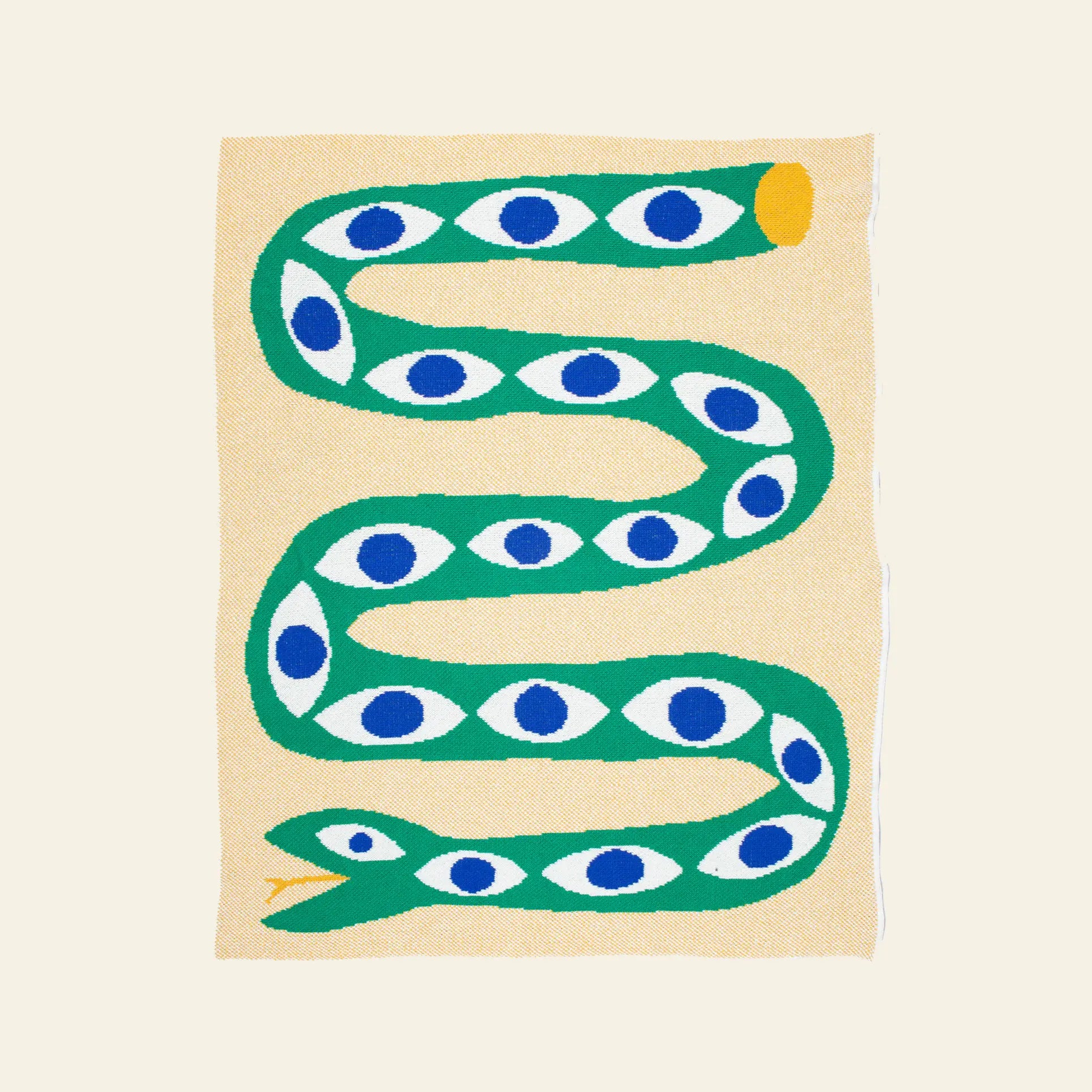The Slowdown Studio Snake Eyes Mini Blanket is a beige throw featuring a green S-shaped snake with blue and white eye patterns, a yellow-tipped tail, and made from eco-friendly recycled cotton.