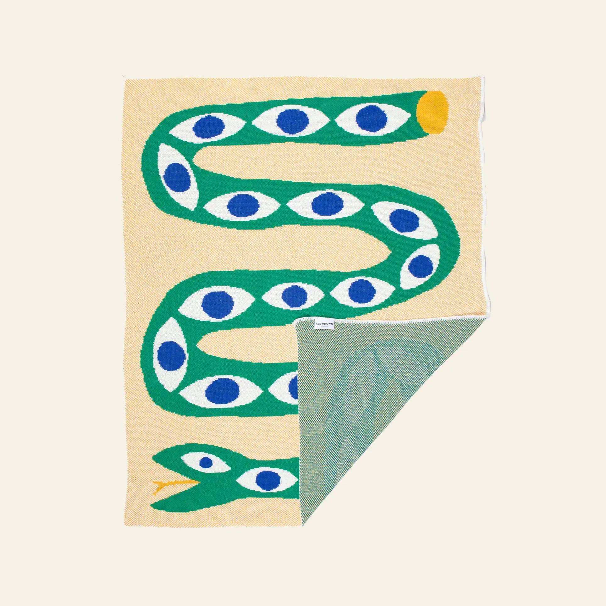 The Slowdown Studio Snake Eyes Mini Blanket, made from recycled cotton, features a beige base with a green snake-inspired design and blue-white eye accents, plus a foldable corner revealing an eco-friendly green underside.