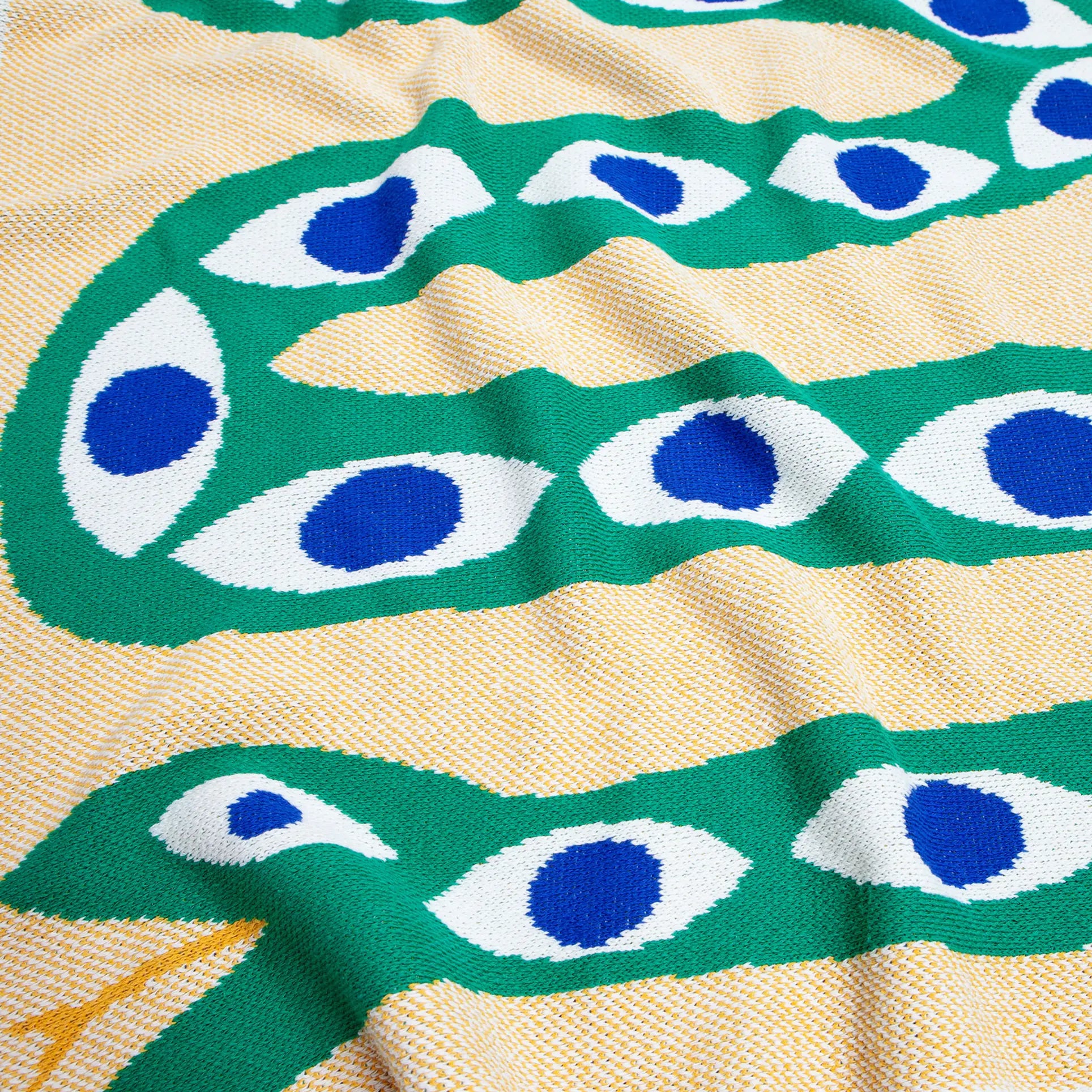 The Slowdown Studio Snake Eyes Mini Blanket by Slowdown Studio showcases a green, snake-inspired pattern with blue and white eye accents on light yellow, crafted from recycled cotton for an eco-friendly touch with a softly wrinkled texture.