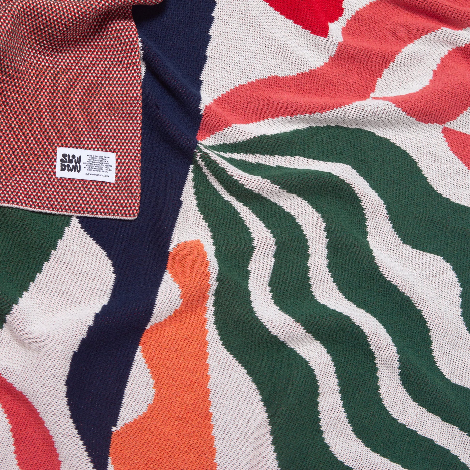 The Slowdown Studio Thurston Knit Blanket by Slowdown Studio is a sustainable, colorful knit made from recycled cotton, with wavy green, orange, red, cream, and navy patterns and a black SLOW DOWN label for cozy comfort.