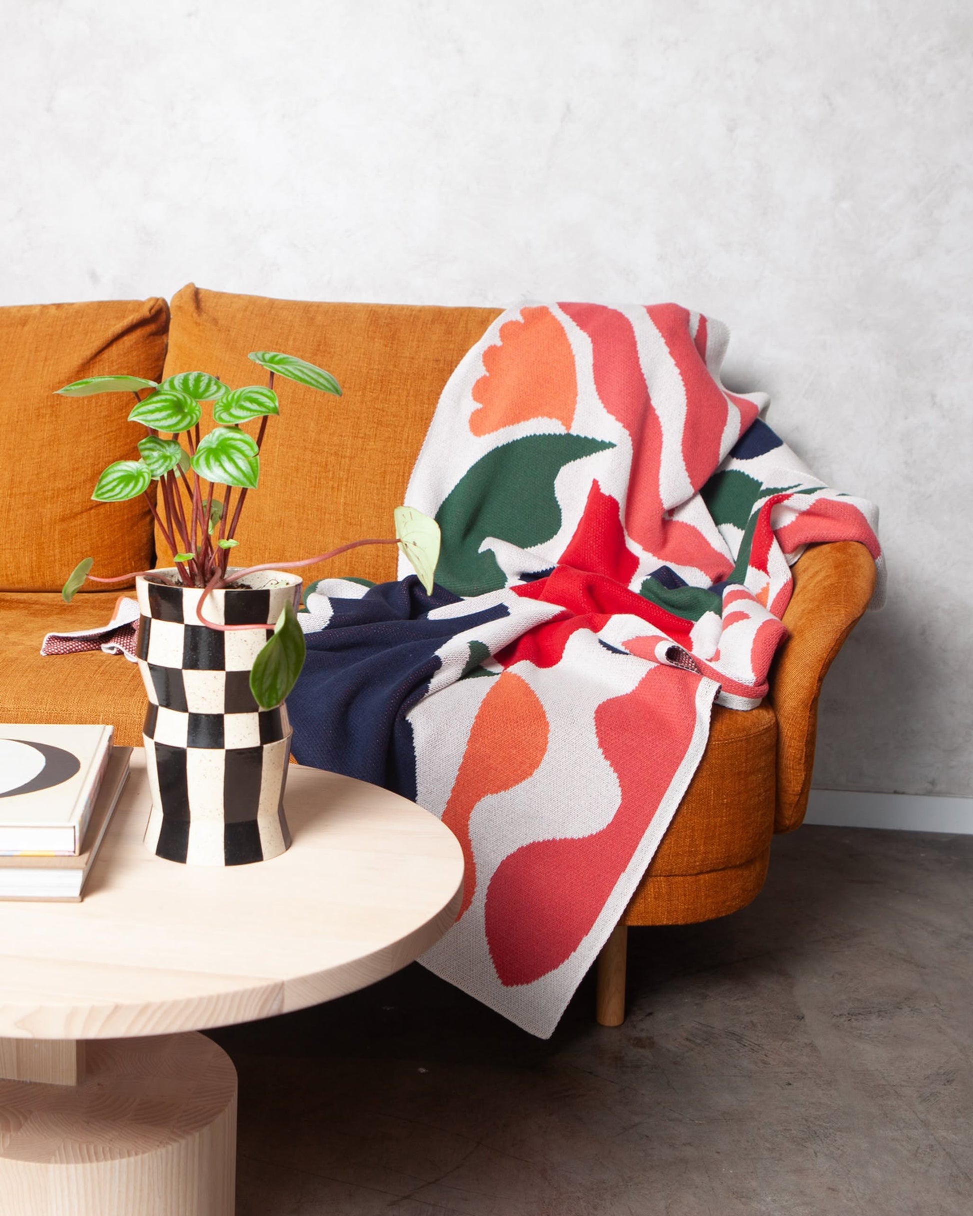 The Slowdown Studio Thurston Knit Blanket, crafted from recycled cotton, is draped over a mustard yellow sofa beside a round wooden table with a checkered vase and green plant in a minimalist, modern space with neutral walls and a concrete floor.