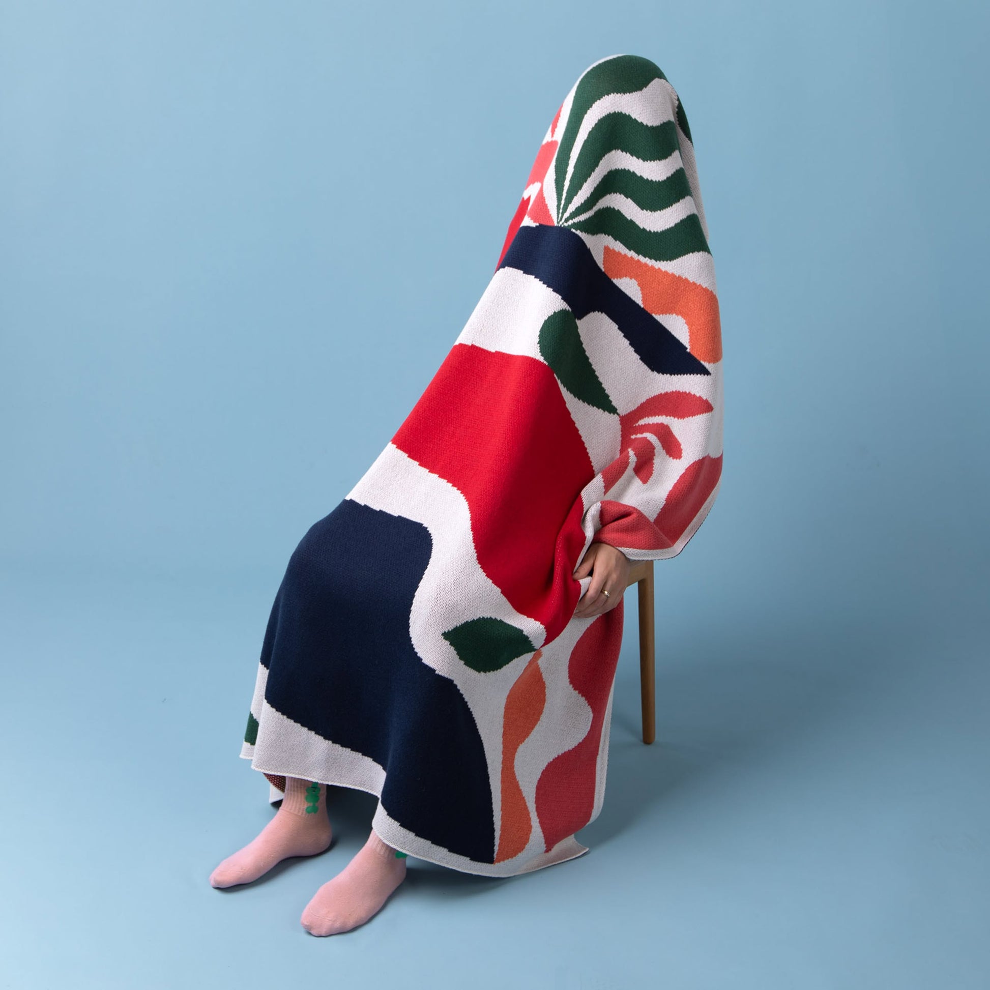 A person sits on a chair against a blue background, covered by the Slowdown Studio Thurston Knit Blanket—an abstract, colorful design in red, navy, green, orange, and white—made from recycled cotton by Slowdown Studio. Only their bare feet are visible.