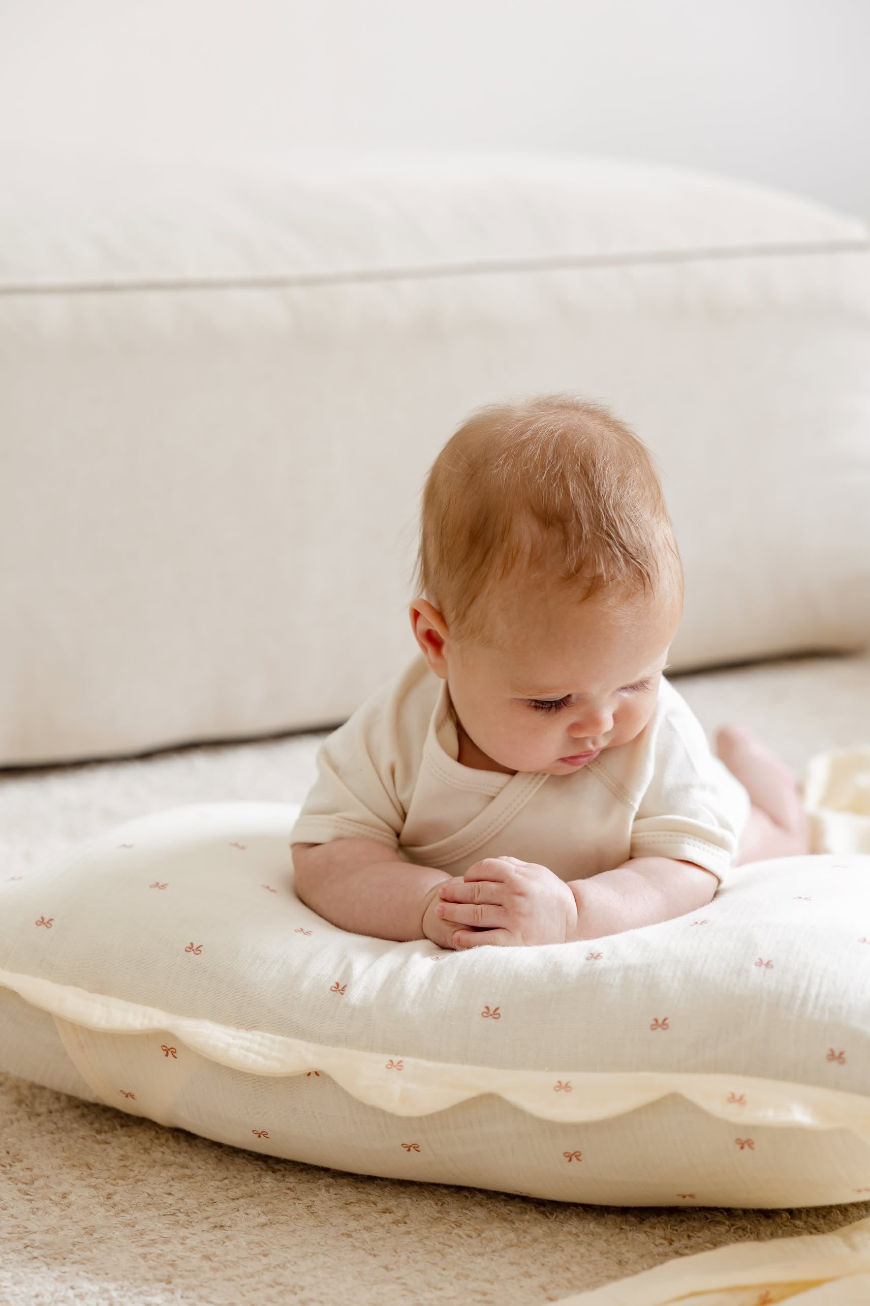 A baby with light brown hair lies on a Soft Spot Soft Petal Cushion Cover (Rectangle, Set of 2) in organic cotton, wearing a cream outfit. The scene includes a pale pillow, plush carpet, and beige sofa in the background.