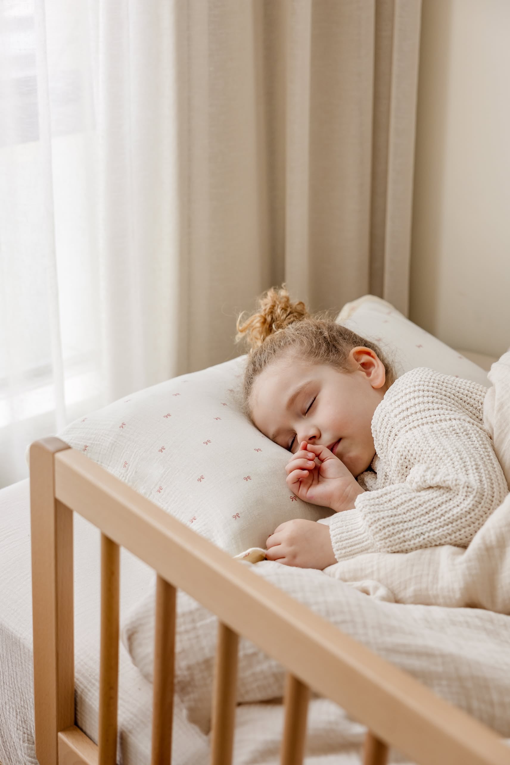 A young child with curly hair sleeps peacefully in a cozy bed, surrounded by Soft Spot Soft Petal Cushion Covers Rectangle, Set of 2, in cream, near a window with sheer curtains.