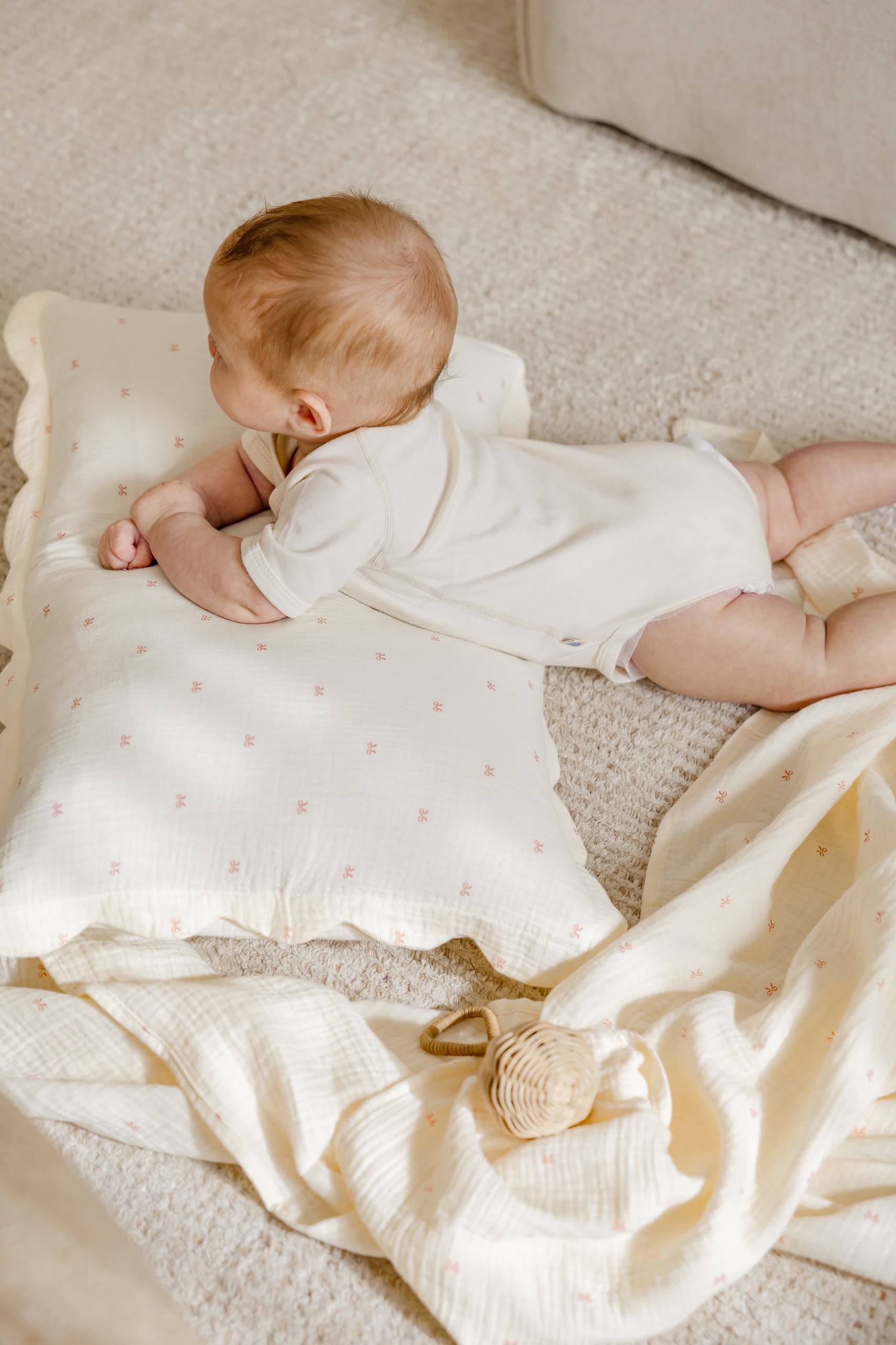 A baby in a cream onesie lies on a soft carpet, resting on Soft Spot Soft Petal Cushion Covers Rectangle, Set of 2 by Soft Spot. A cream blanket and small woven rattle are nearby.