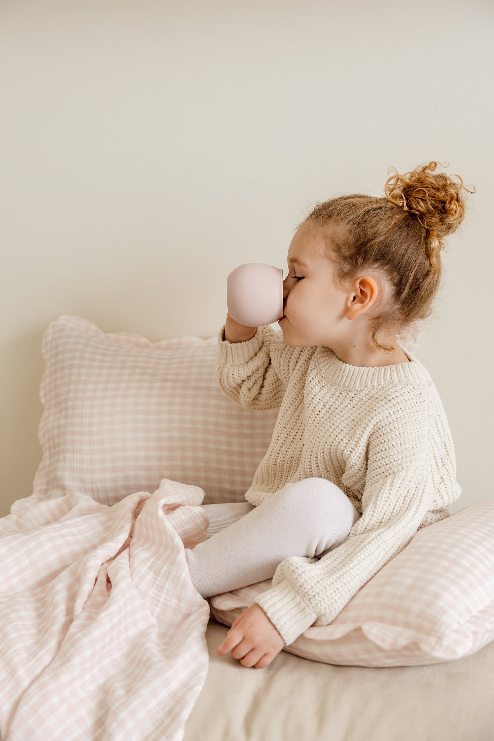 A young child with curly hair sits on a bed with Soft Spot Soft Petal Cushion Covers Rectangle, Set of 2, wearing a cream sweater and white leggings, drinking from a light pink cup.