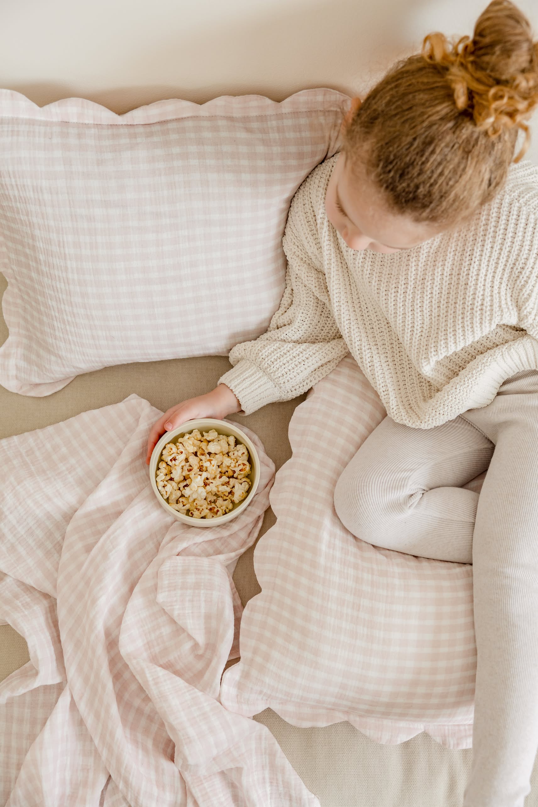 A young girl with curly hair in a bun sits on a bed with pink plaid bedding, holding popcorn. She wears a beige sweater and light leggings atop Soft Spot Soft Petal Cushion Covers Rectangle, Set of 2 by Soft Spot.