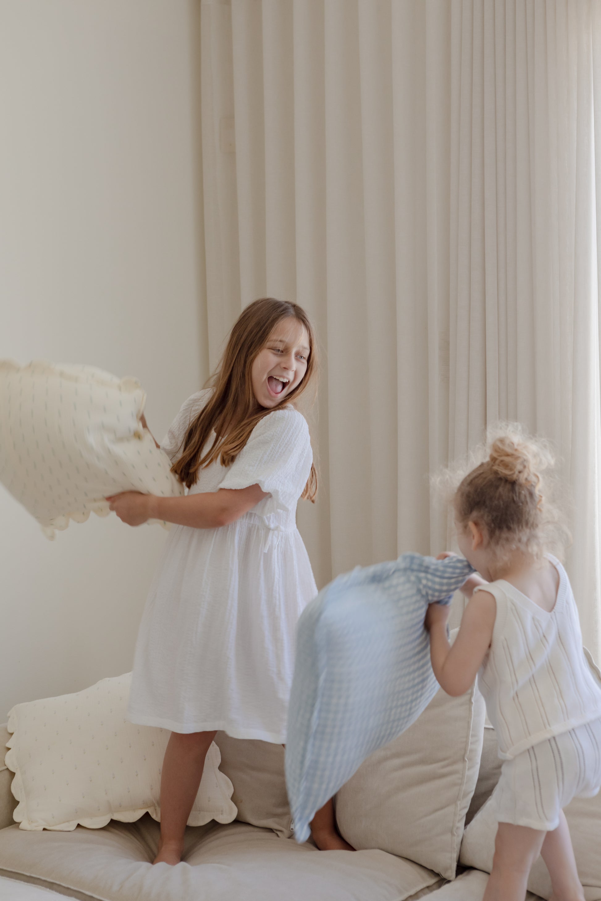 Two young girls laugh and toss Soft Spot Soft Petal Cushion Covers, Set of 2, made from light-colored fabric, on a beige sofa in a bright room with cream-colored curtains.