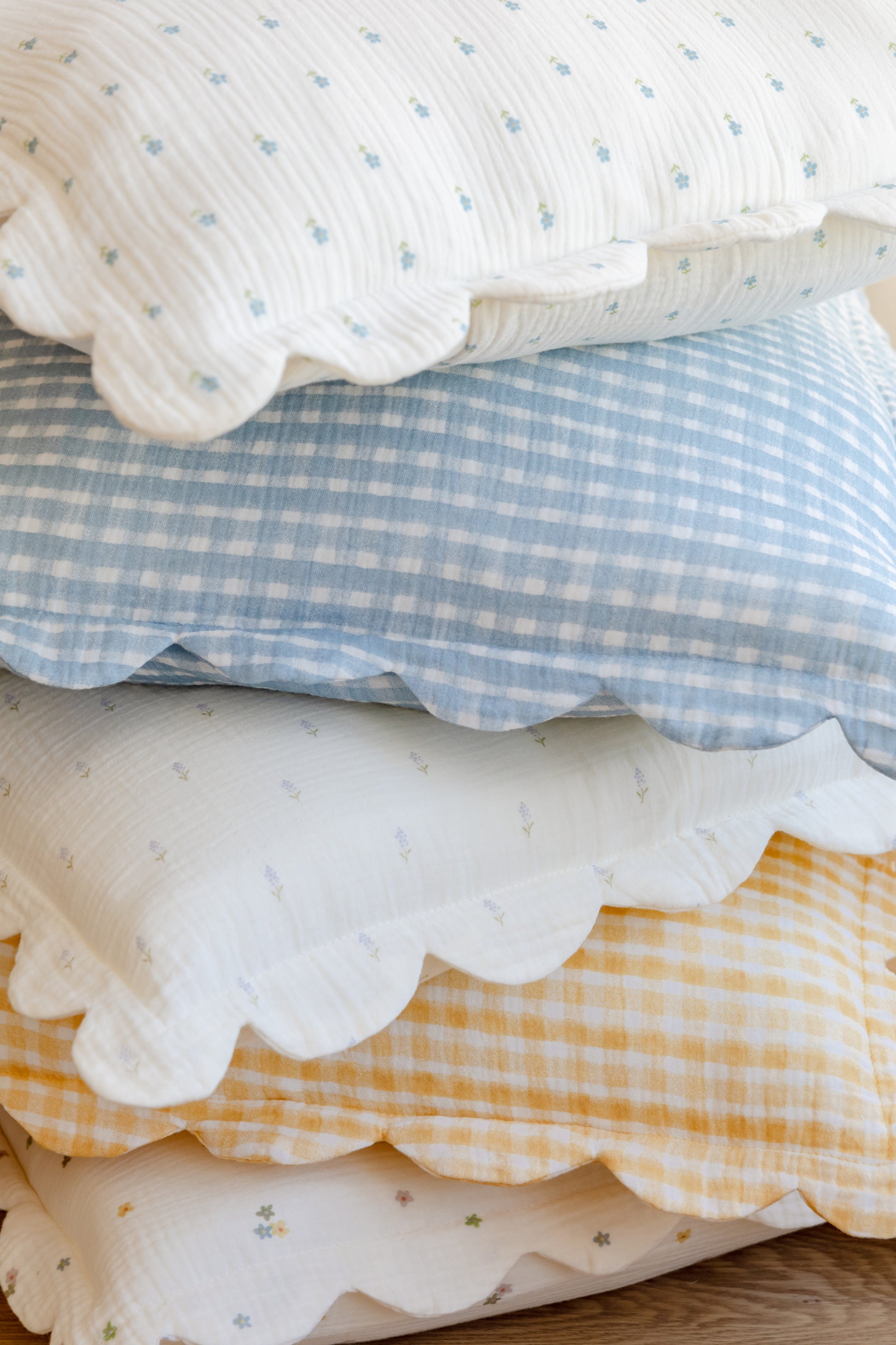 A stack of two Soft Spot Soft Petal Cushion Covers, featuring pastel florals and gingham patterns, is neatly arranged on a wooden surface.