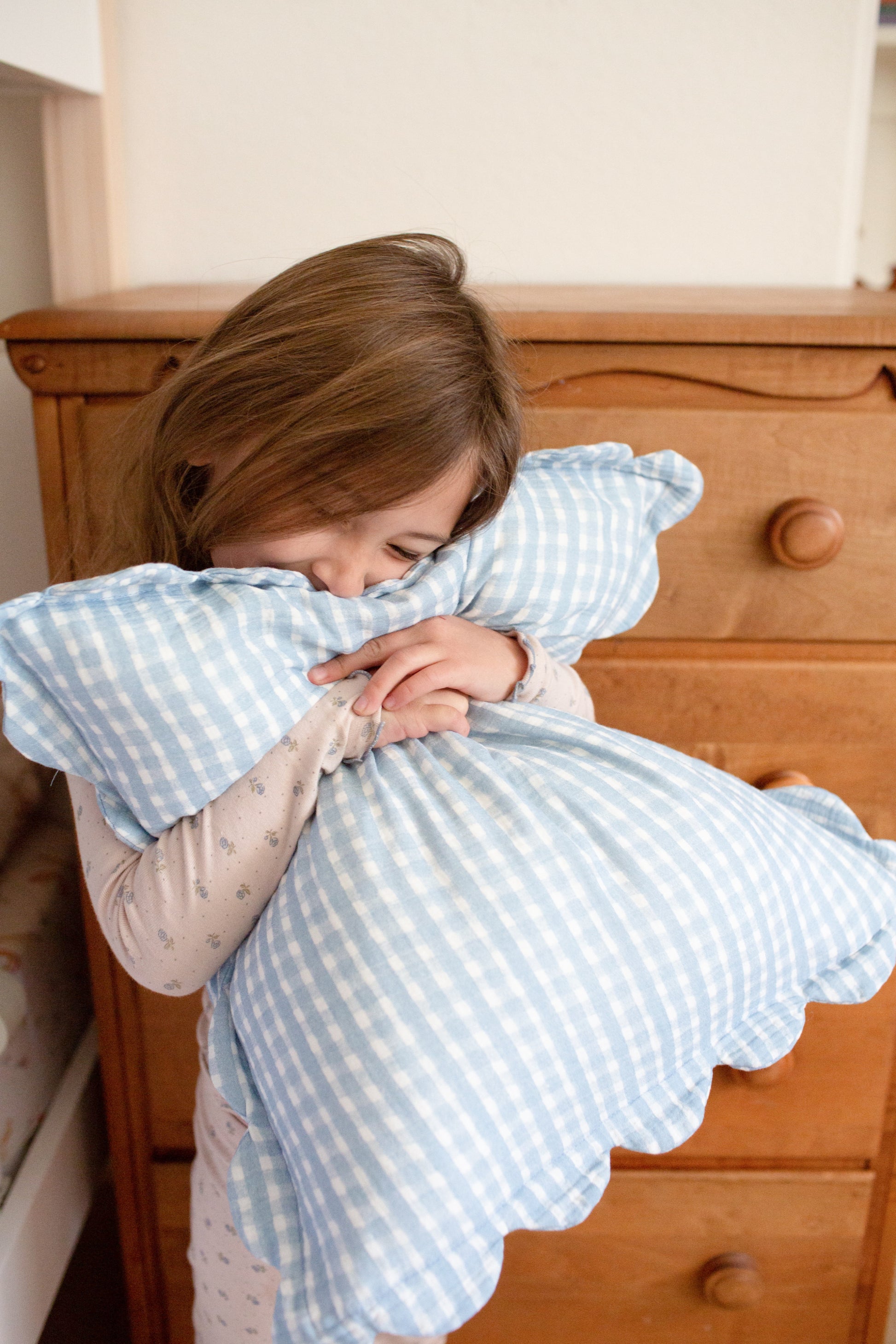 A young child with brown hair hugs a blue and white Soft Spot Soft Petal Cushion Cover from the set of 2 by Soft Spot, smiling with eyes closed while standing in front of a wooden dresser in a cozy bedroom.