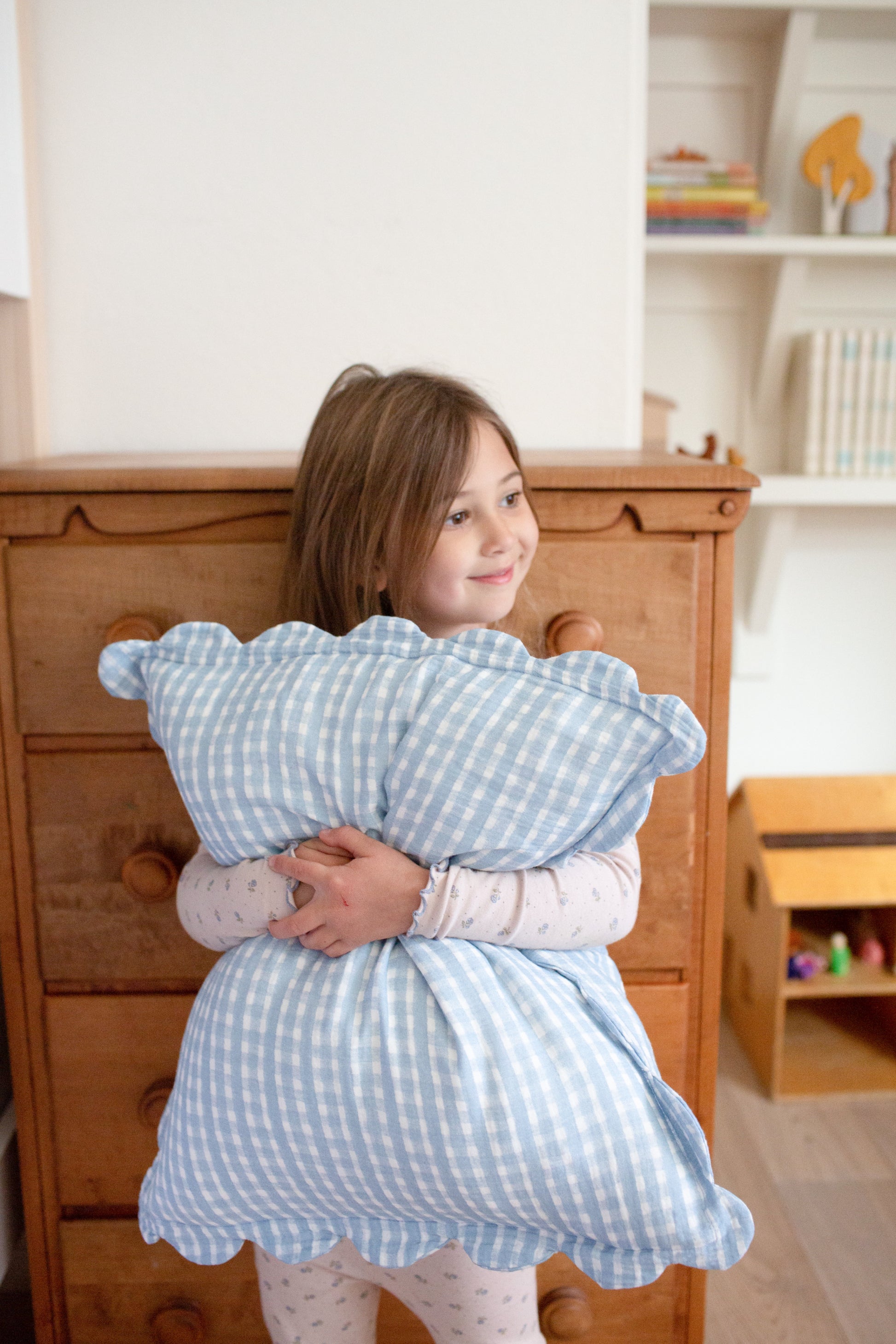 A young girl with light brown hair in pajamas smiles softly while hugging a large pillow covered with Soft Spot Soft Petal Cushion Covers, Set of 2—featuring blue-and-white checks and scalloped edges—in a cozy room with a wooden dresser and shelves behind her.