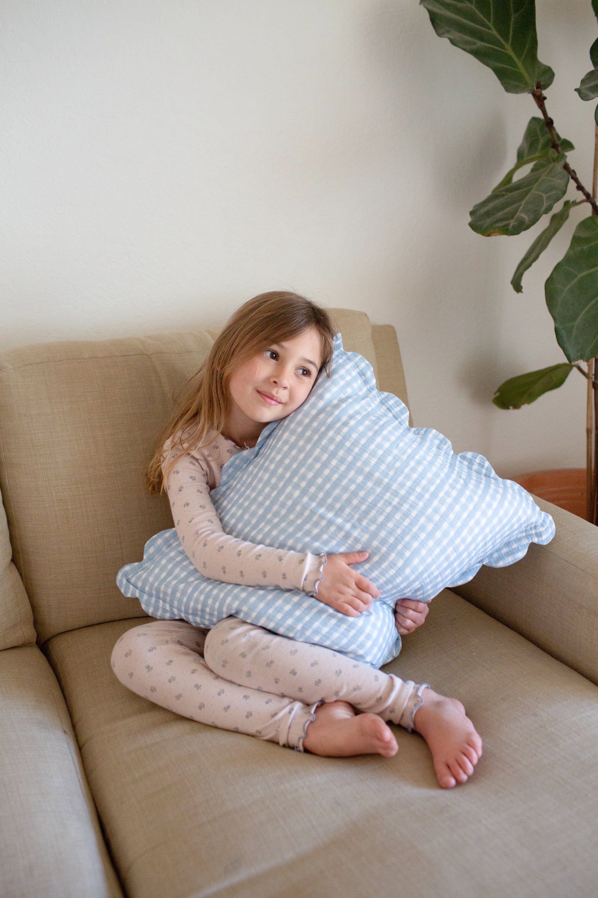 A young girl in light pajamas hugs a blue and white Soft Spot Soft Petal Cushion Cover (Set of 2) by Soft Spot on a beige sofa, with a green plant in the background.