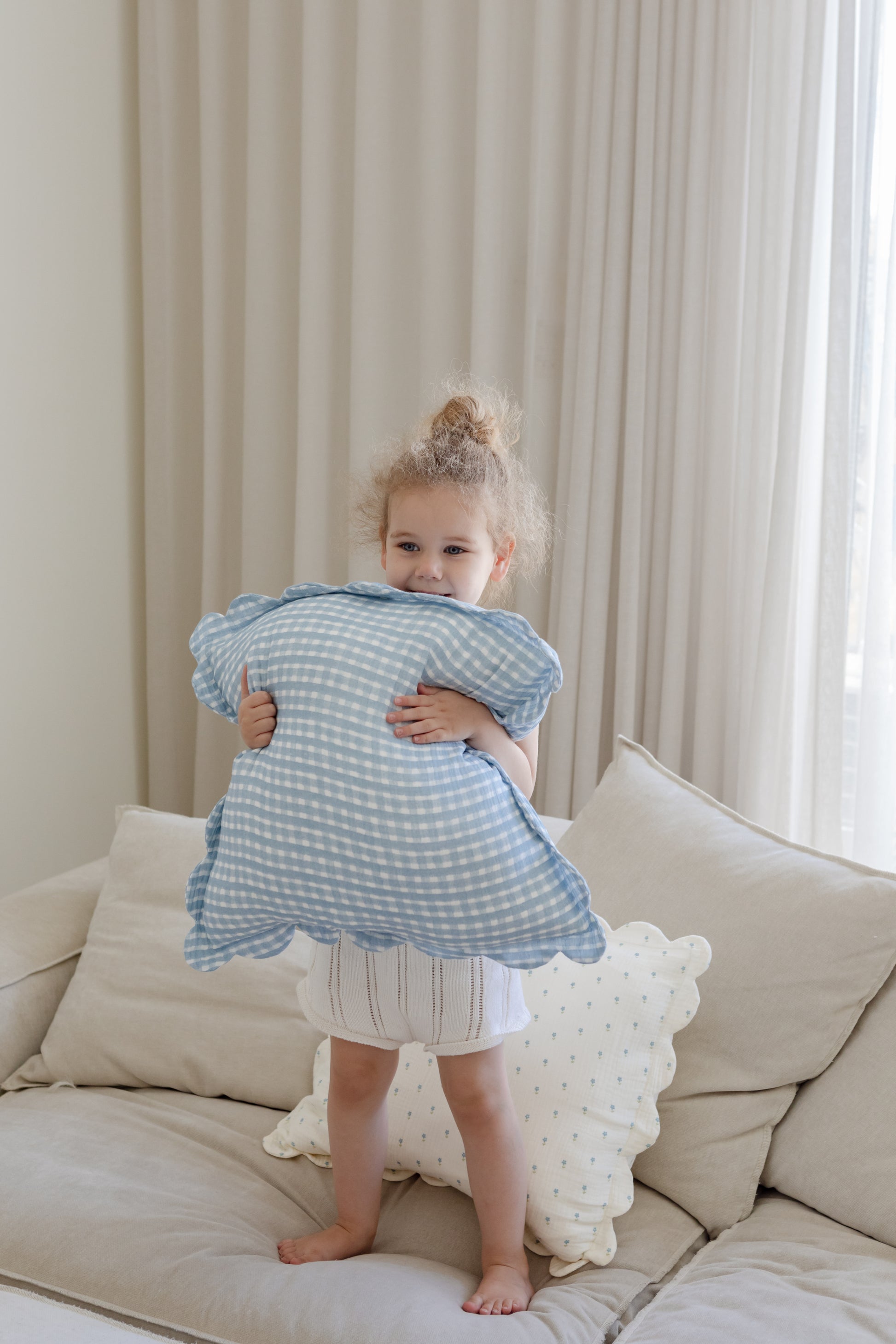 A young child with tied-up curly hair smiles on a beige couch, holding a blue gingham cloud pillow. The cozy scene features Soft Spots Soft Petal Cushion Covers (Set of 2) and light curtains by the window in the background.