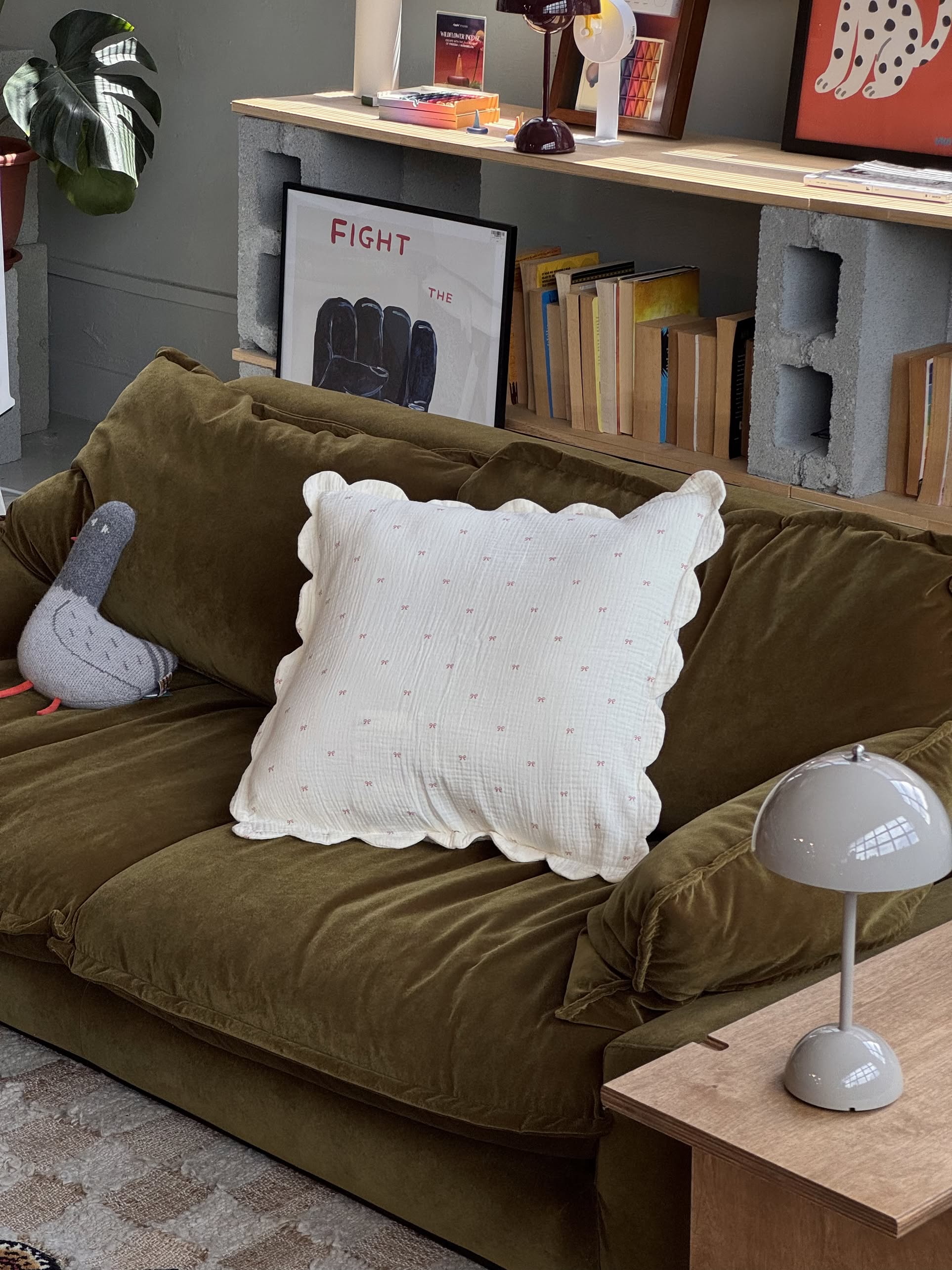 An olive green couch with a large white Soft Spot Soft Petal Cushion Cover and organic cotton, a gray plush toy, and a small table lamp. Behind the couch are shelves with books, art, and a FIGHT THE poster. Brand: Soft Spot.