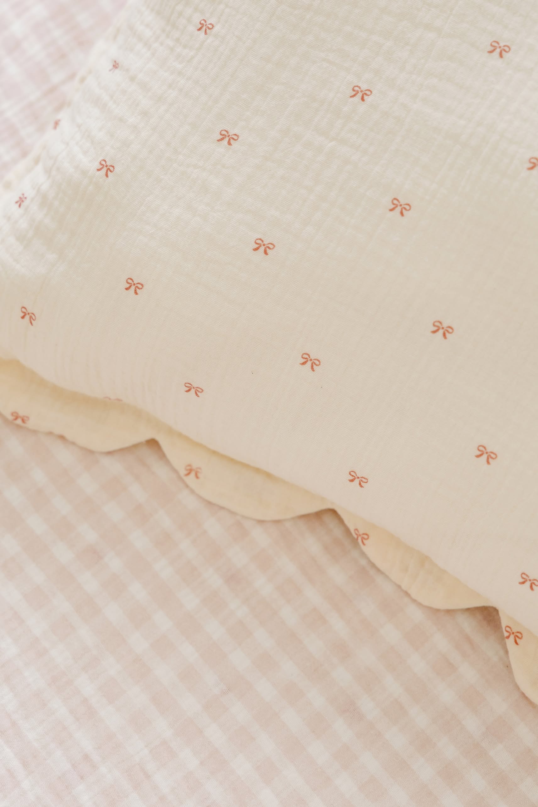 A close-up of Soft Spot’s Soft Petal Cushion Covers, set of 2, in cream with pink bows, shown on a pale pink and white gingham sheet.