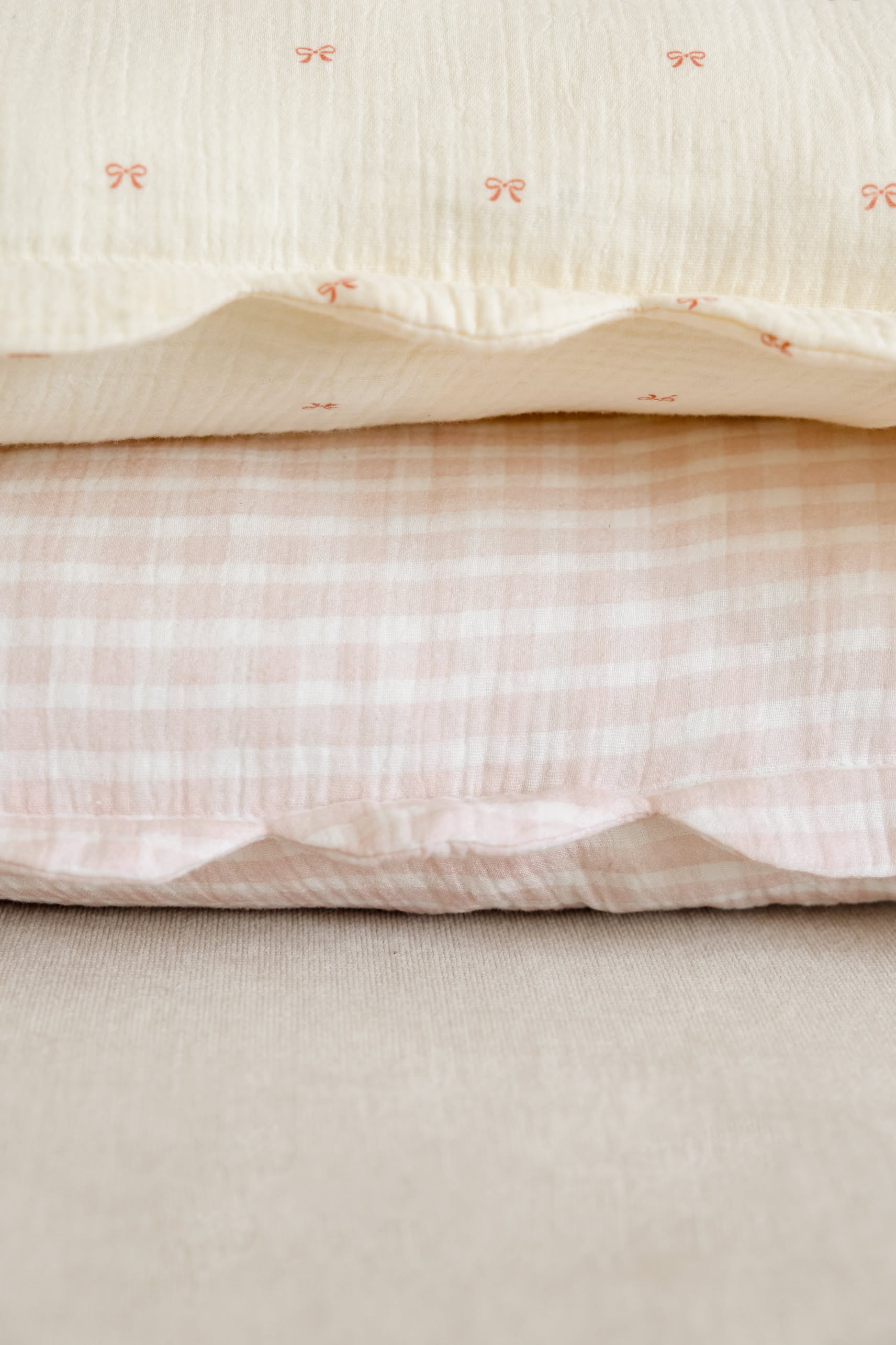 The Soft Spot Soft Petal Cushion Covers, Set of 2 by Soft Spot, includes a cream cover with red bows and a pink-and-white gingham check, both crafted from organic cotton for extra comfort.