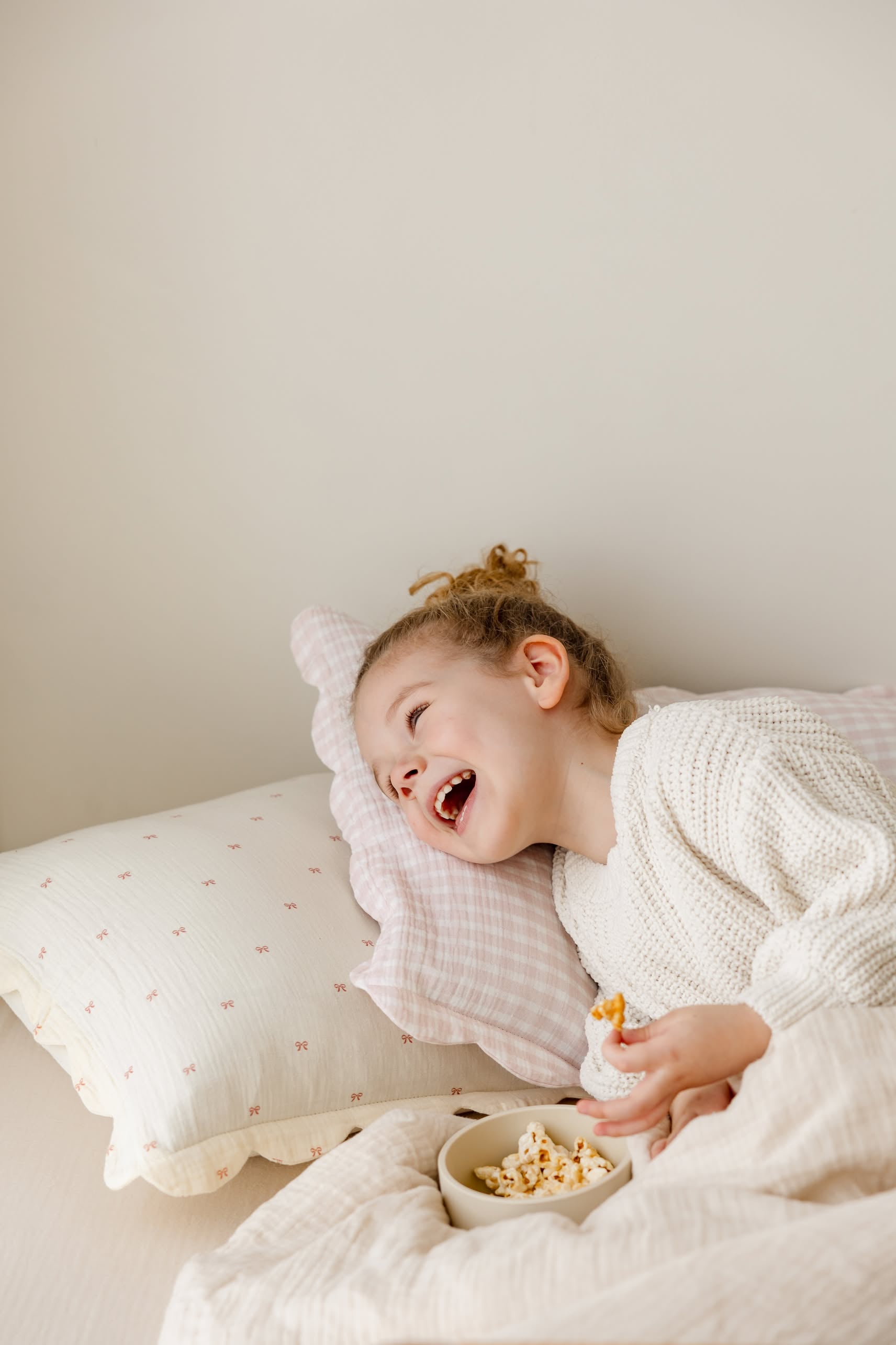 A young child with curly hair, wearing a white sweater, smiles and laughs on a bed with pale pillows and Soft Spot Soft Petal Cushion Covers, Set of 2 by Soft Spot, while holding a bowl of snacks.