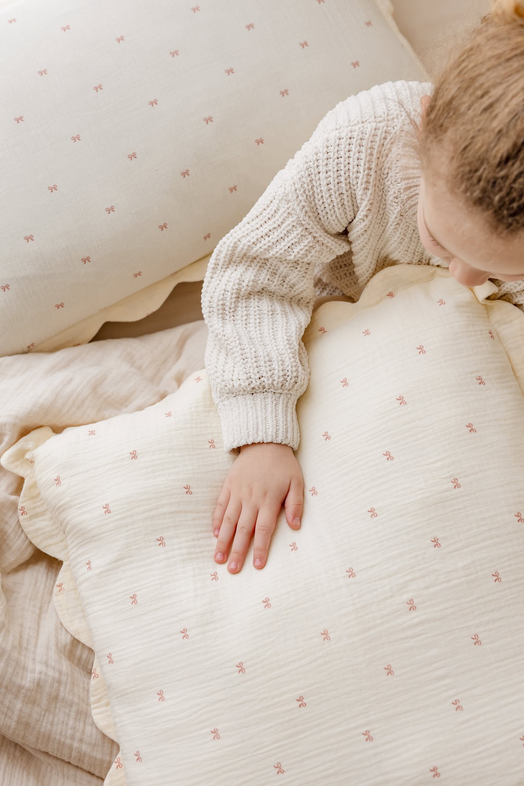 A young child in a knitted sweater rests on cream bedding with tiny pink patterns, surrounded by Soft Spot’s Soft Petal Cushion Covers (Set of 2) and matching bedding.