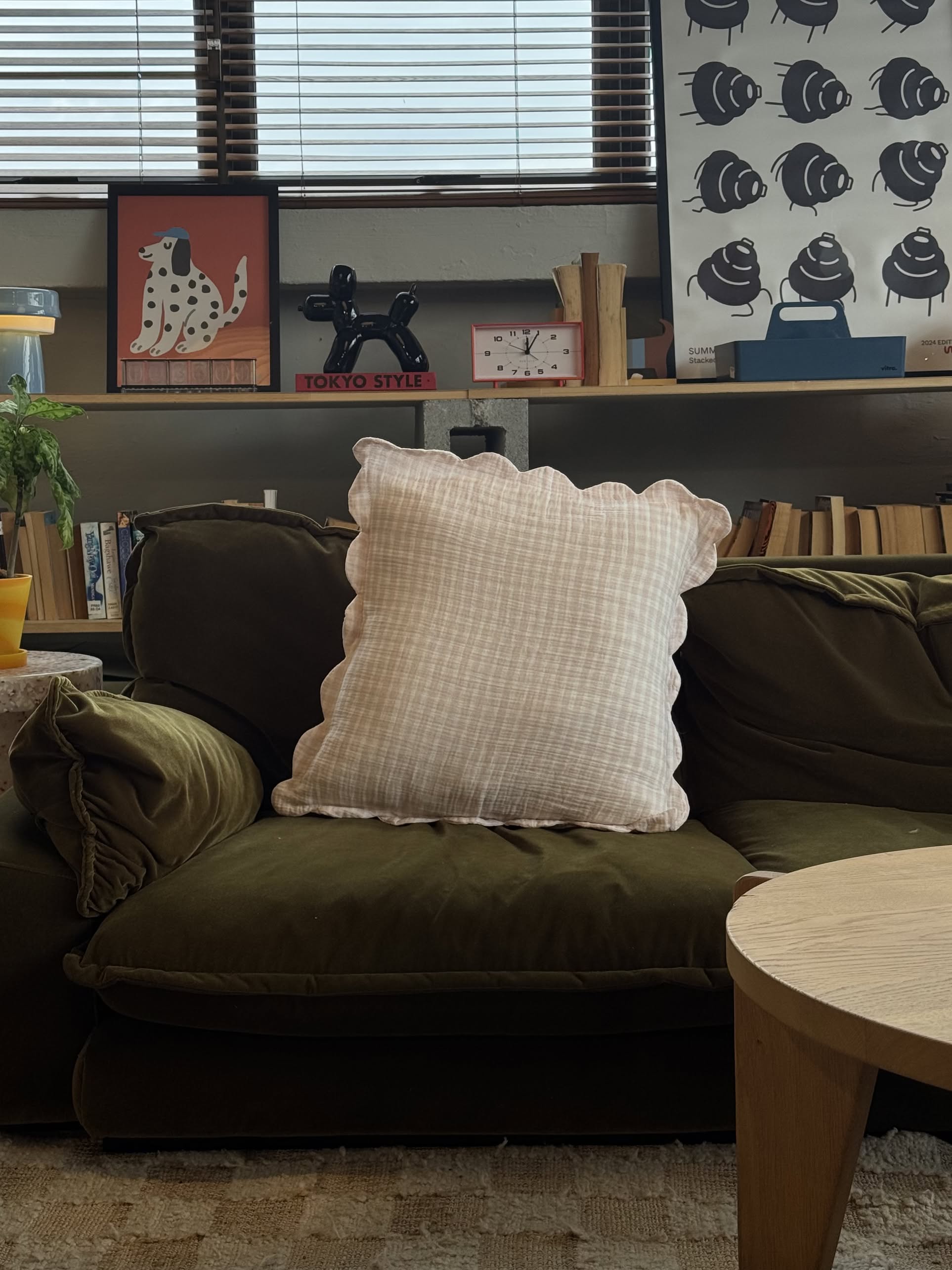 A green velvet couch features a white, scalloped-edge pillow and Soft Spot Soft Petal Cushion Covers, Set of 2. In front is a low bookshelf with books and art, plus a small table with a gray lamp. Gentle daylight fills the inviting space.