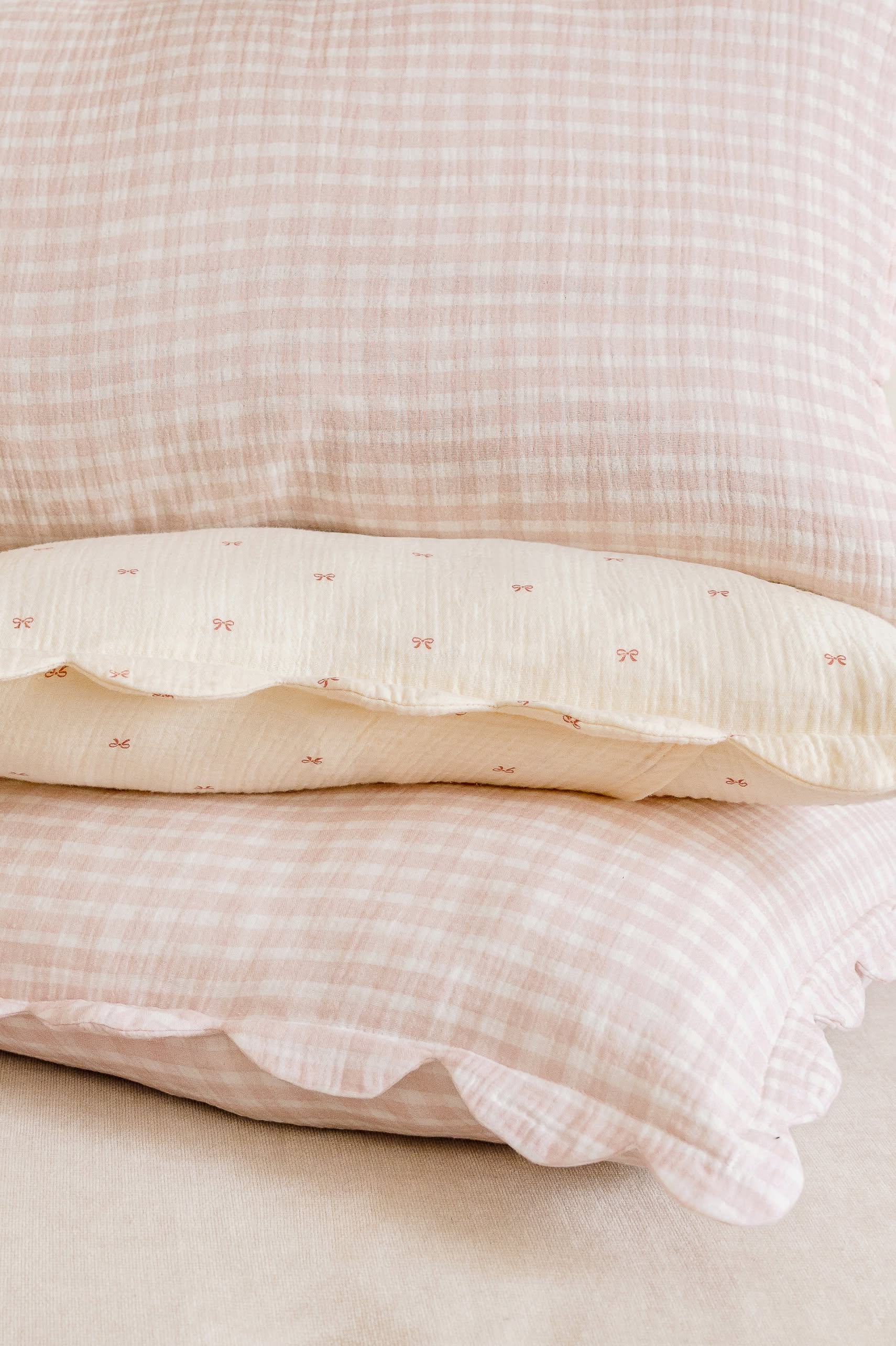 A close-up of two stacked Soft Spot Soft Petal Cushion Covers in pale pink plaid with envelope closures and small bow patterns, plus a cream pillow between them, all by Soft Spot, creates a cozy and delicate bedding look.