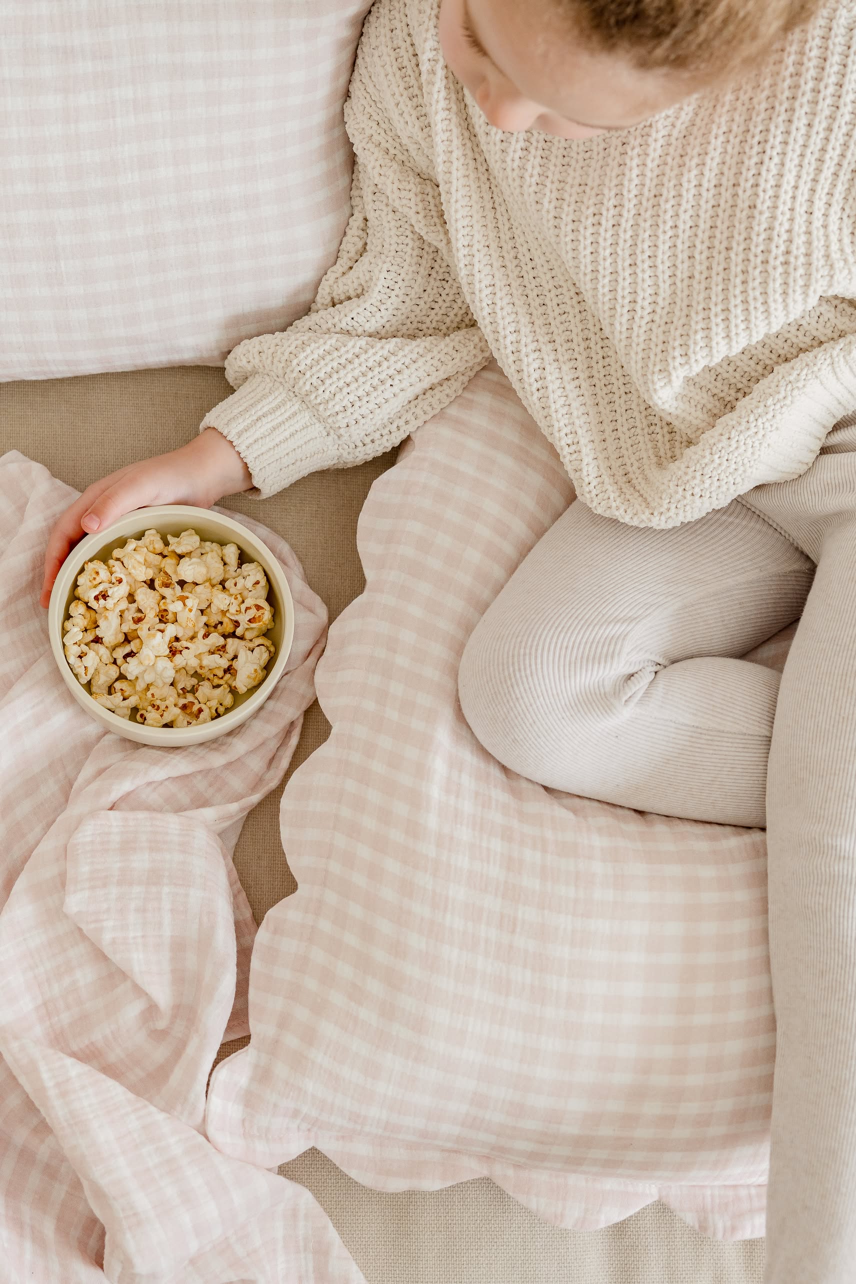 Wearing a cozy cream sweater and light leggings, a person sits on a couch with Soft Spot Soft Petal Cushion Covers, Set of 2, and pink plaid pillows, holding a bowl of popcorn.