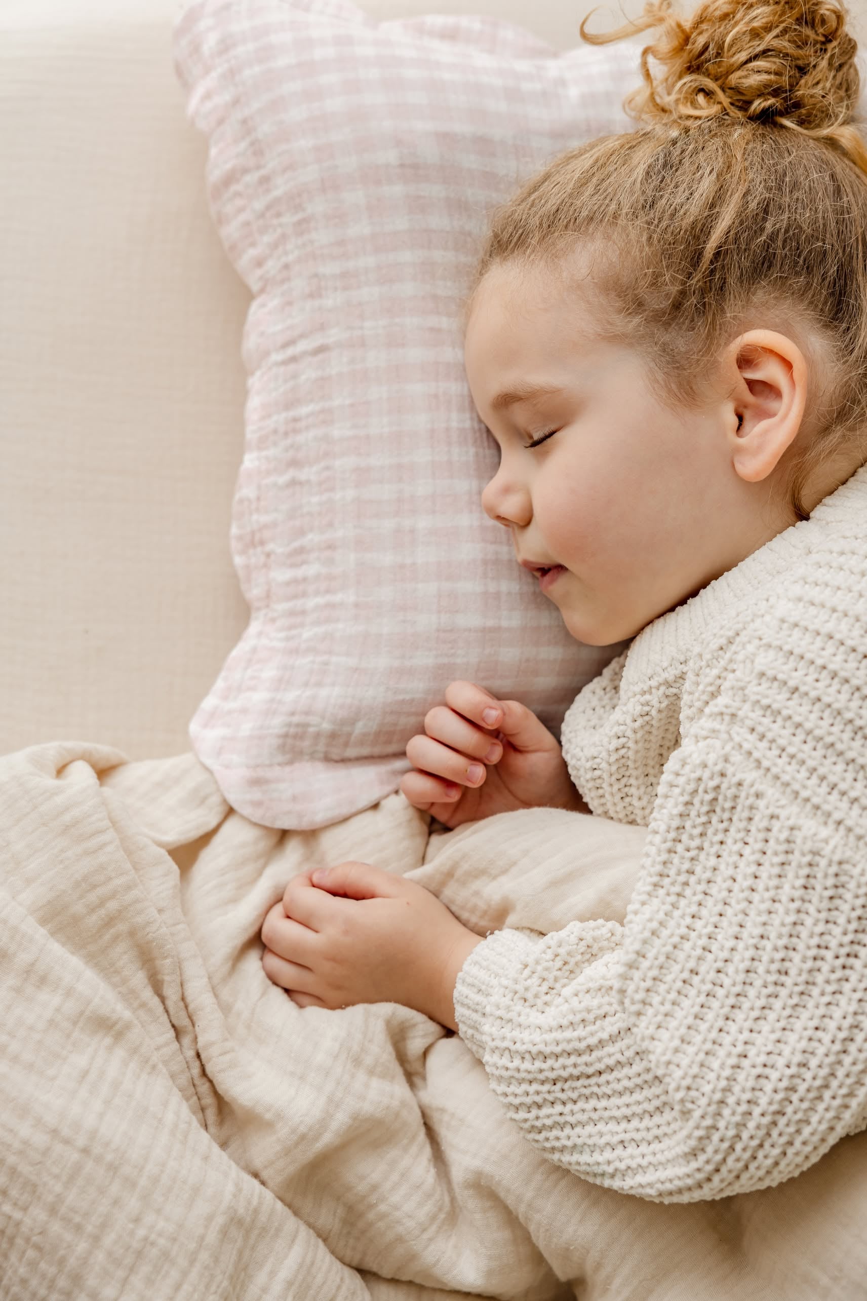 A young child with curly hair sleeps peacefully on a soft bed, covered with a cream blanket and resting on a pink plaid pillow from the Soft Spot Soft Petal Cushion Covers, Set of 2 by Soft Spot.