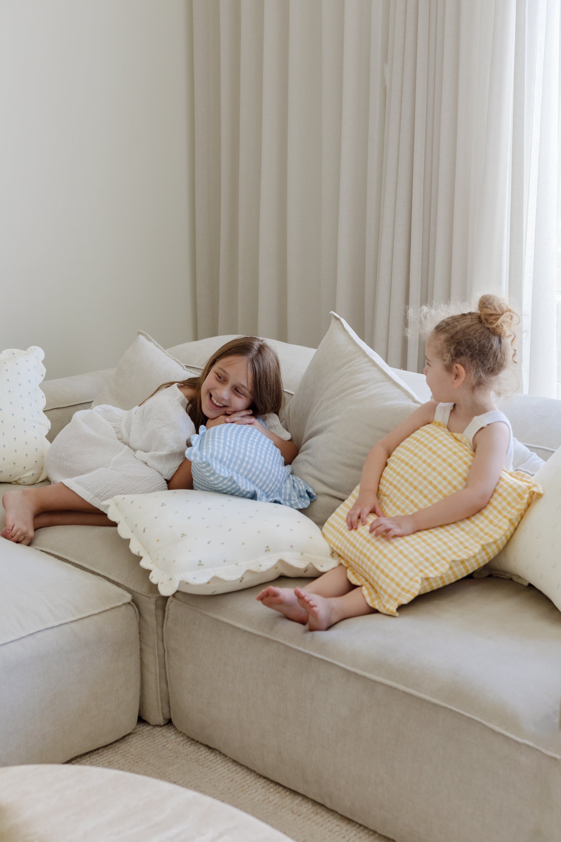 Two young girls smile on a light-colored sectional, holding Soft Spot Soft Petal Cushion Covers (Set of 2). Soft lighting and sheer curtains complete the cozy, relaxed atmosphere.
