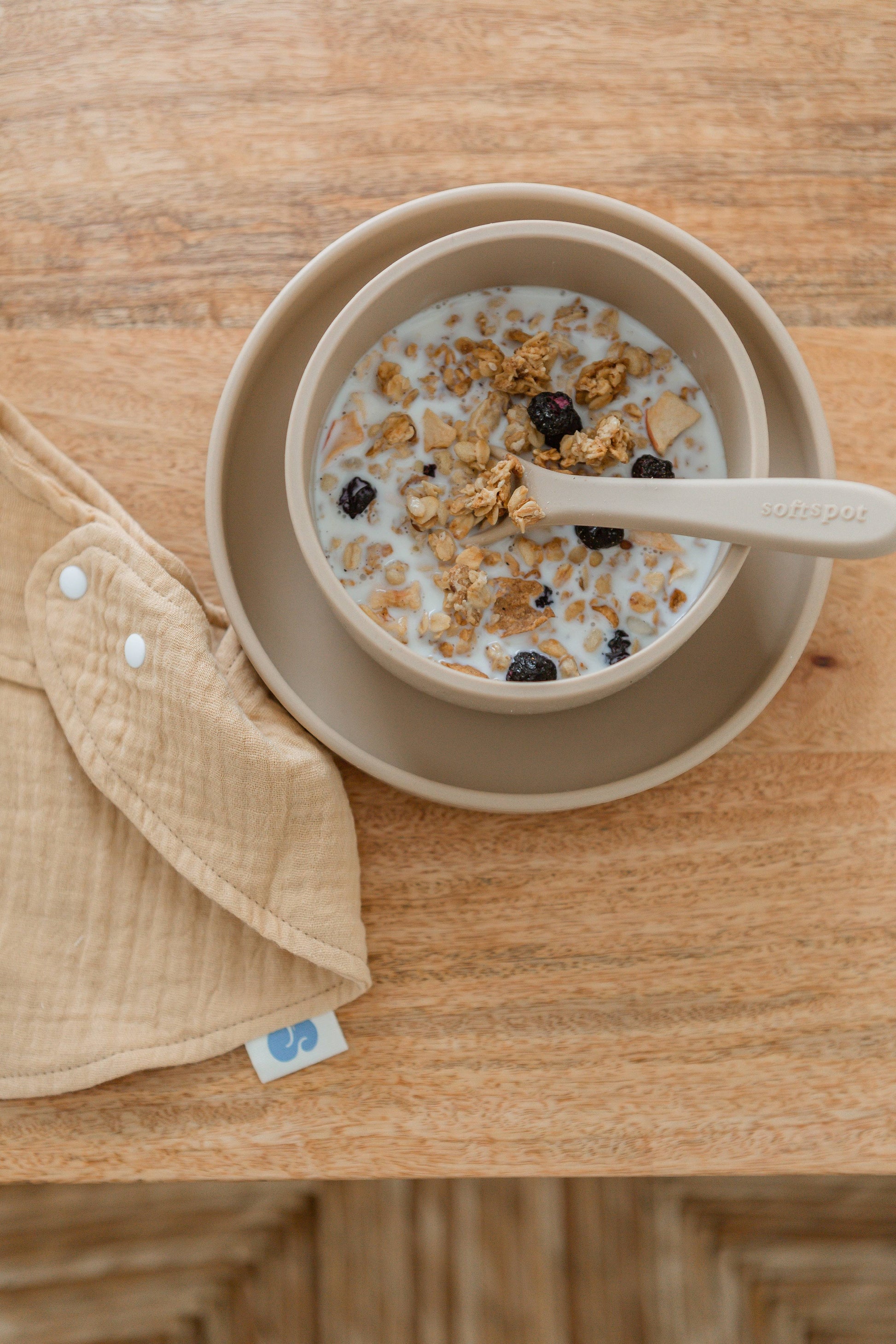The Soft Spot Soft Suction Bowl by Soft Spot in beige, filled with cereal, granola, and dried berries, sits on a matching plate with a beige spoon. Its suction grip keeps it steady on the wooden table next to a beige cloth with white snaps.