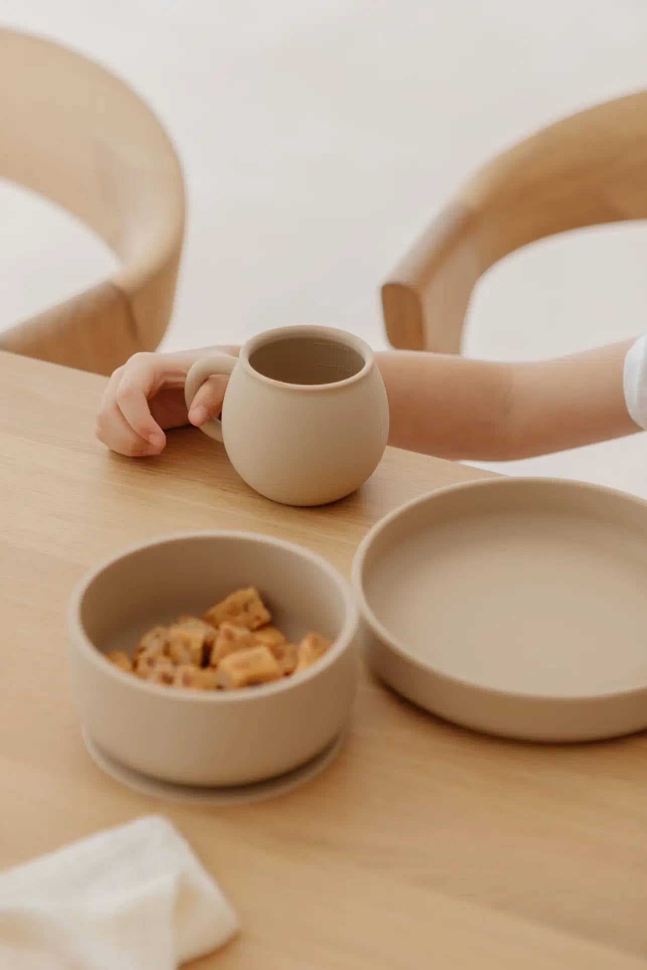 A child’s hand holds a beige food-grade silicone mug beside a matching plate and Soft Spot Soft Suction Bowls by Soft Spot, filled with snacks on a light wooden table with curved wooden chairs in the background.