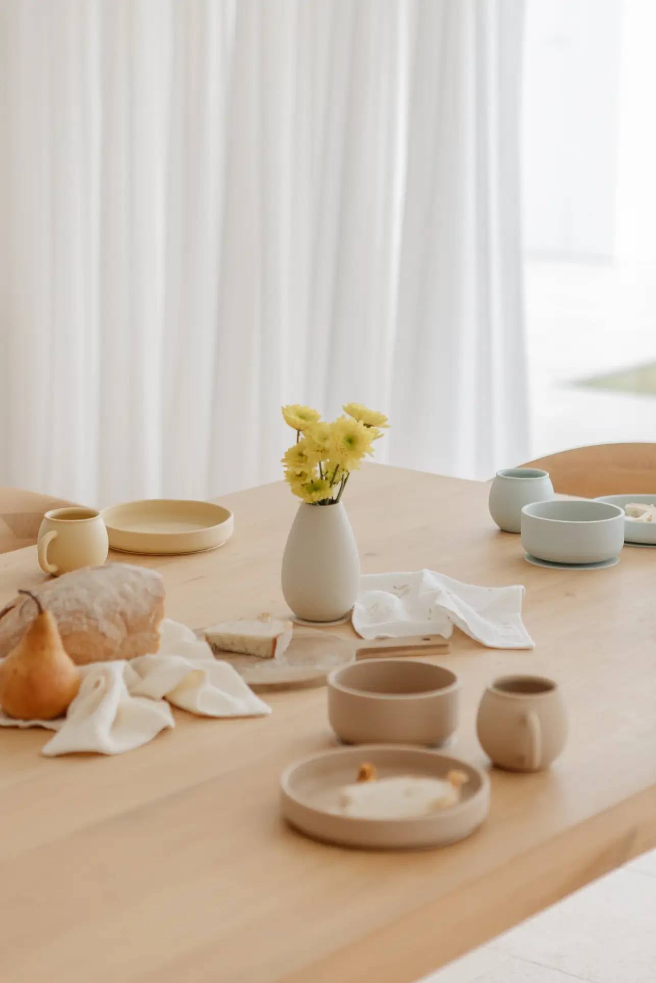 A wooden table features Soft Spot Soft Suction Bowls by Soft Spot (food-grade silicone), ceramic dishes, a mug, bread, a pear, and yellow flowers in a vase. White cloth napkins are scattered with sheer white curtains in the background.