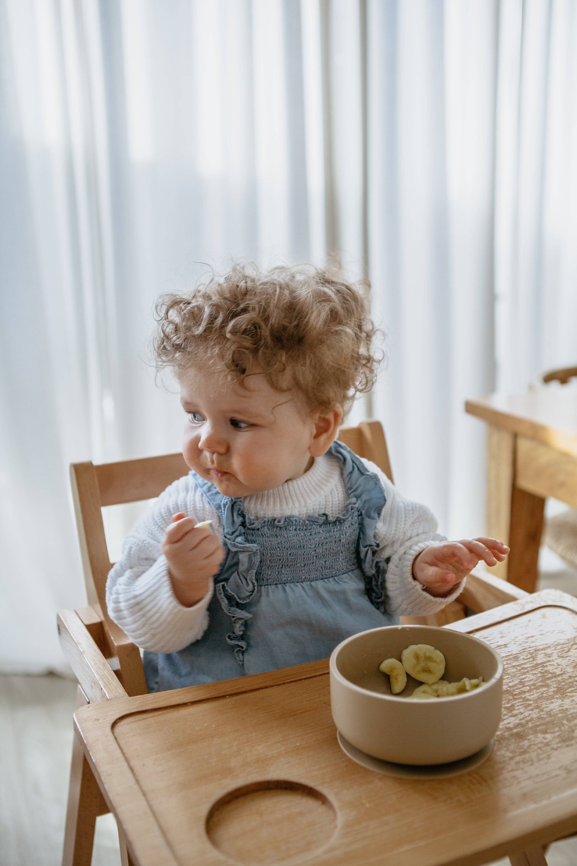 A toddler with curly hair, in a white sweater and blue overalls, sits in a wooden high chair eating pasta from the Soft Spot Soft Suction Bowl, as soft daylight filters through white curtains in the background.