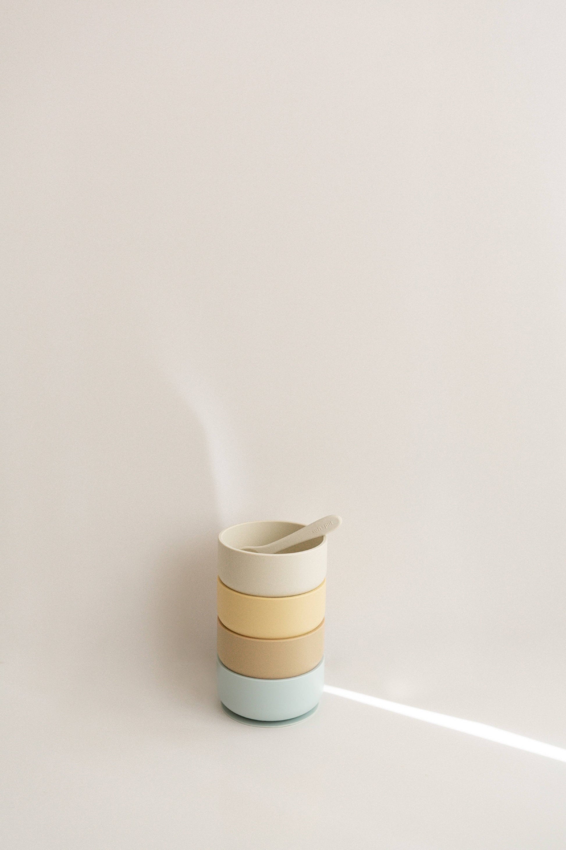 A stack of Soft Spot Soft Suction Bowls in pastel colors, made from food-grade silicone with a spoon inside, sits against a plain, light background under soft lighting.