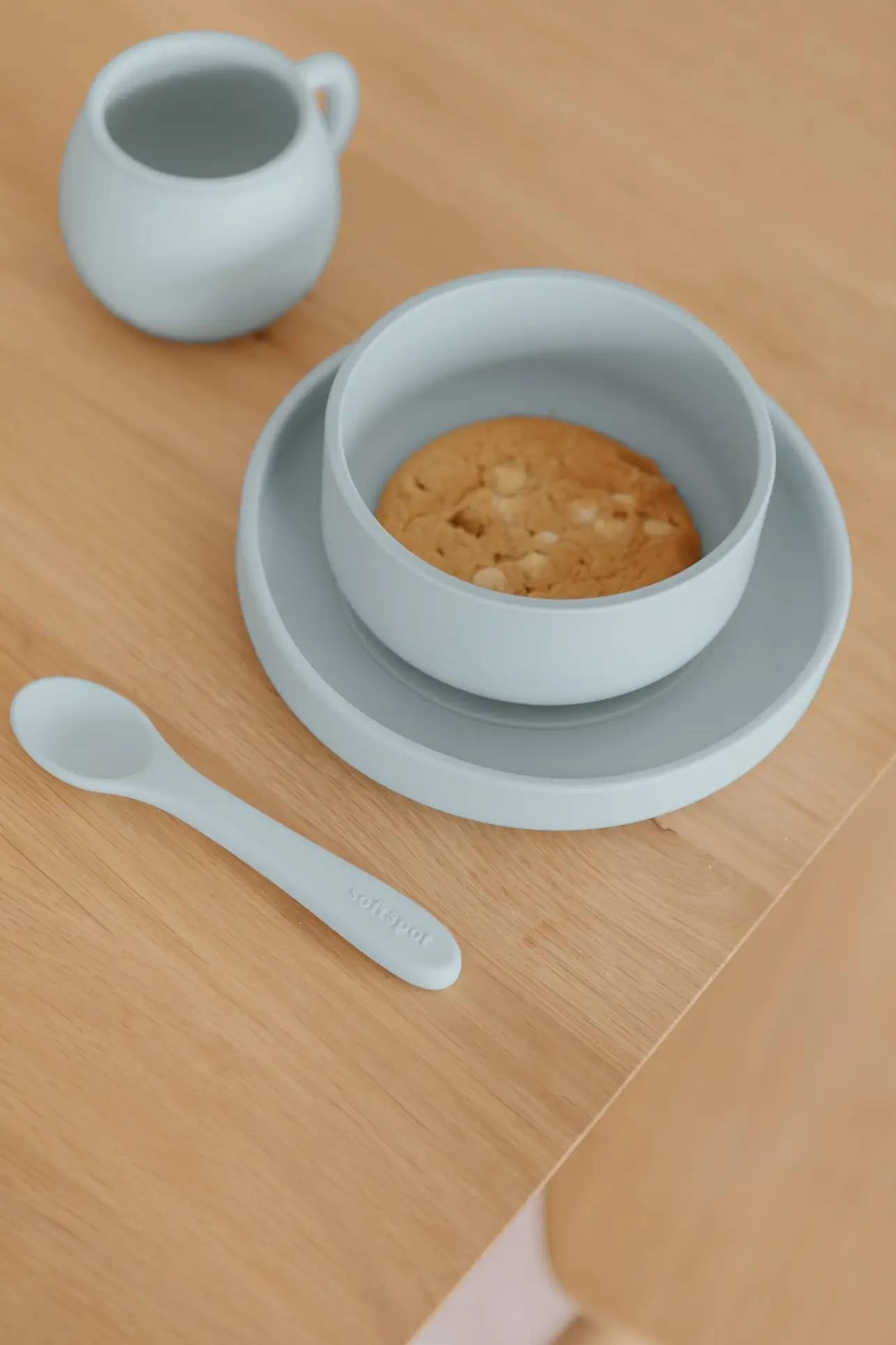 The Soft Spot Soft Suction Bowl by Soft Spot in light blue, made from food-grade silicone, sits on a matching plate with a cookie inside. Complete with suction grip, its paired with a spoon and small pitcher on a light wooden table.